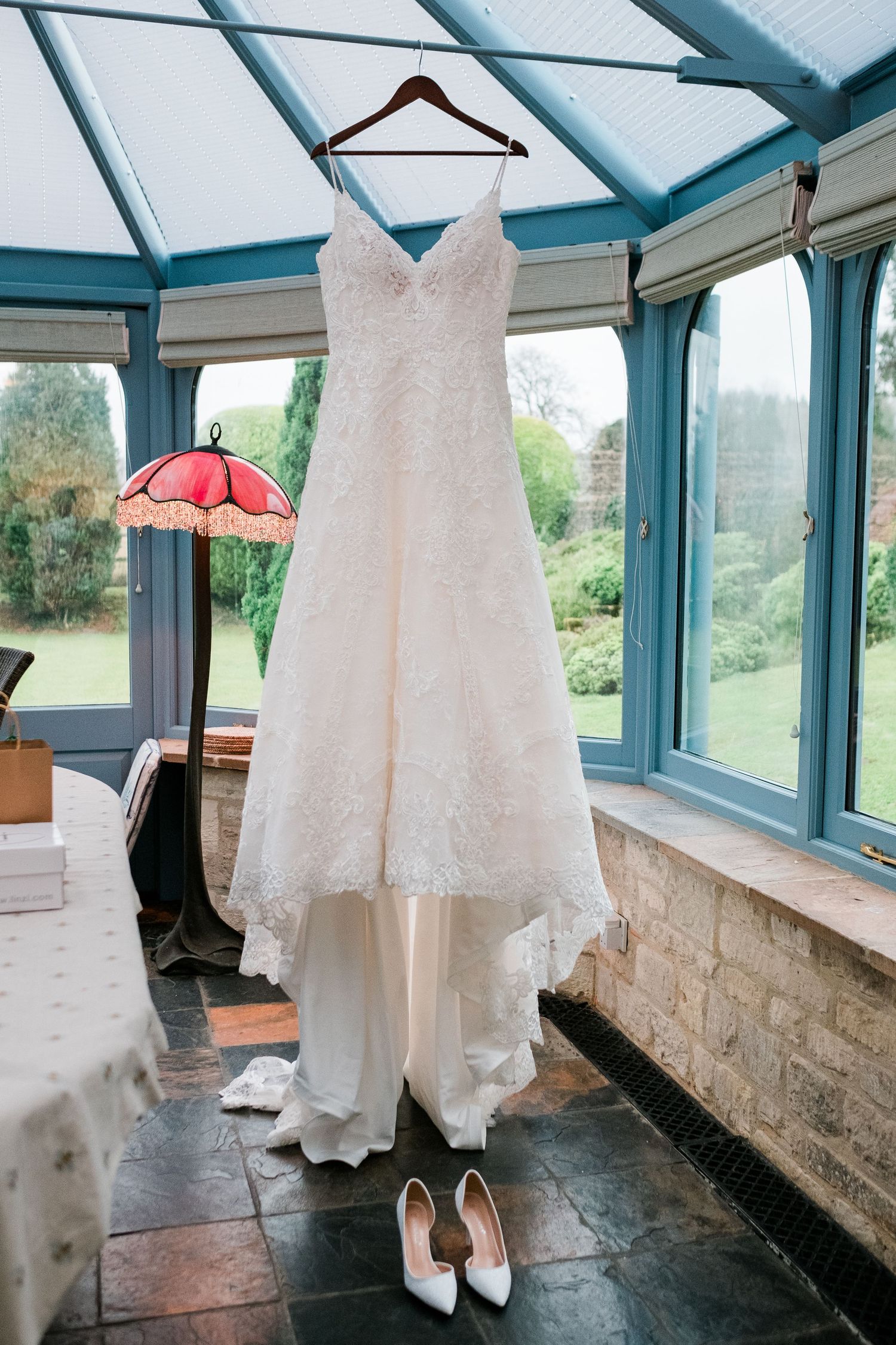 White lace wedding dress and shoes displayed in a bright conservatory with blue-framed windows.