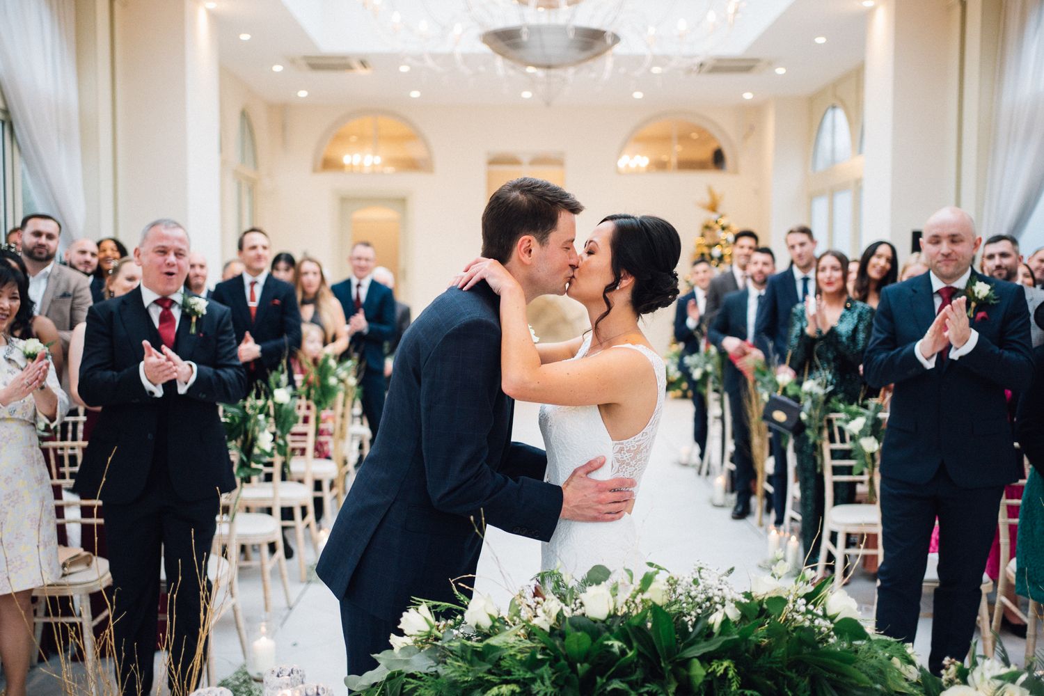 Bride and groom share first kiss at wedding ceremony aisle surrounded by applauding guests in elegant venue.