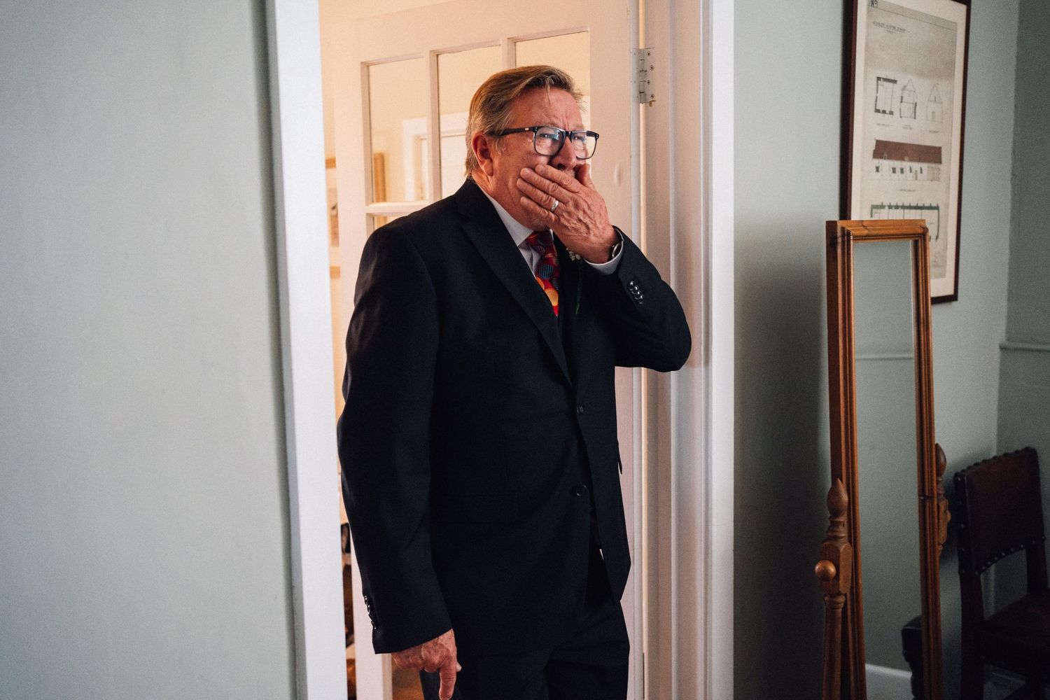 A person in a dark suit and glasses stands in a doorway with their hand covering their mouth.