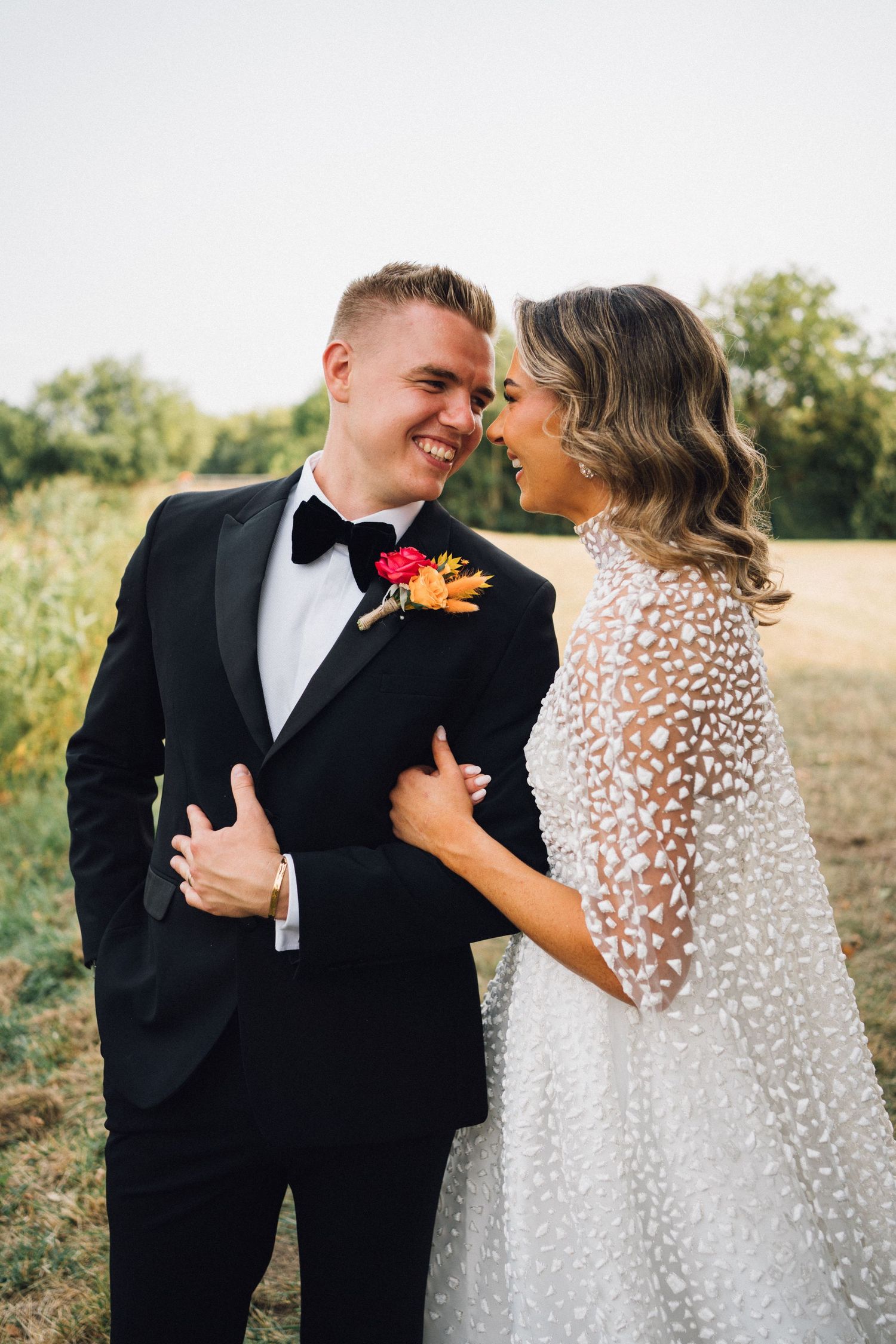 A couple shares intimate moments in a field at sunset, with the bride wearing a polka dot veil and groom in classic tuxedo.
