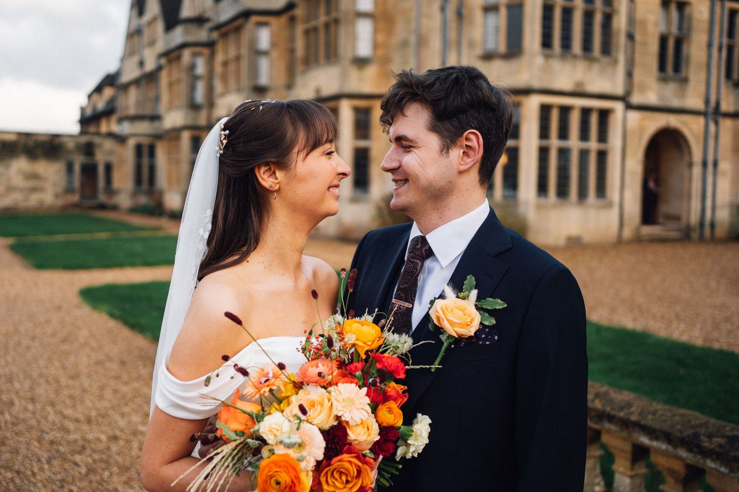 Newlywed couple smiling at each other in front of historic stone building, bride holding colorful bouquet.