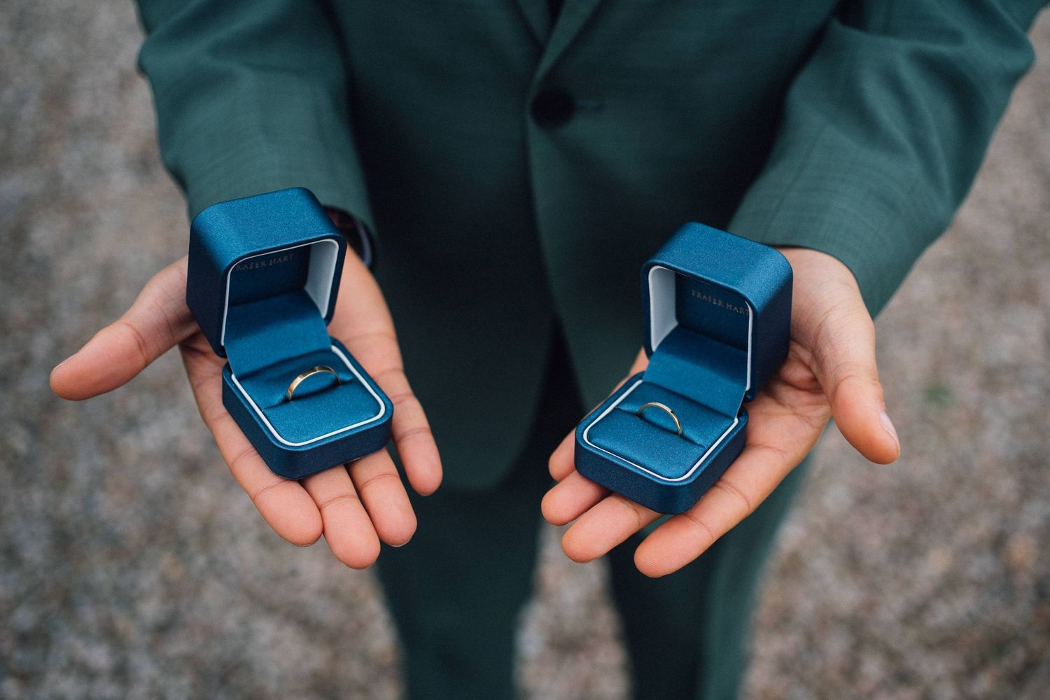 Two blue velvet ring boxes held in outstretched hands wearing a dark green jacket.
