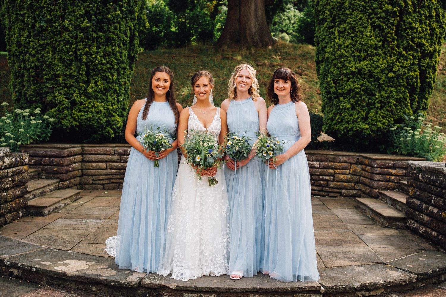 Wedding party members in light blue dresses pose together on stone steps surrounded by greenery.