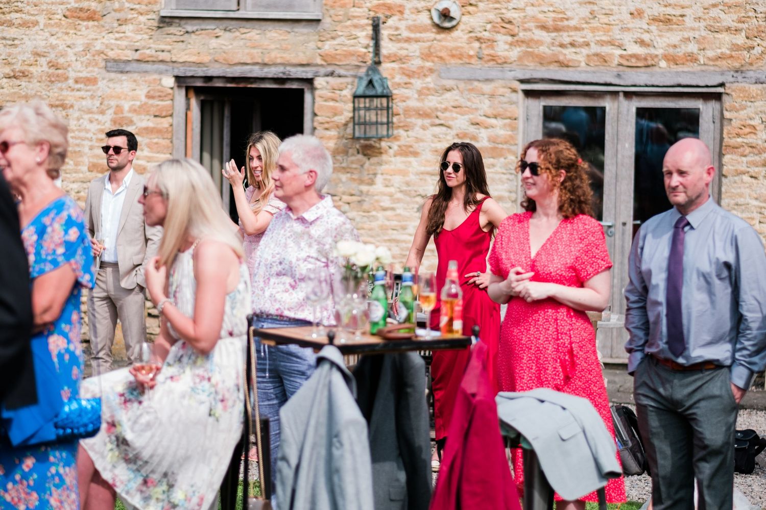 Party guests in colorful summer attire socialize during an outdoor gathering near a stone building.