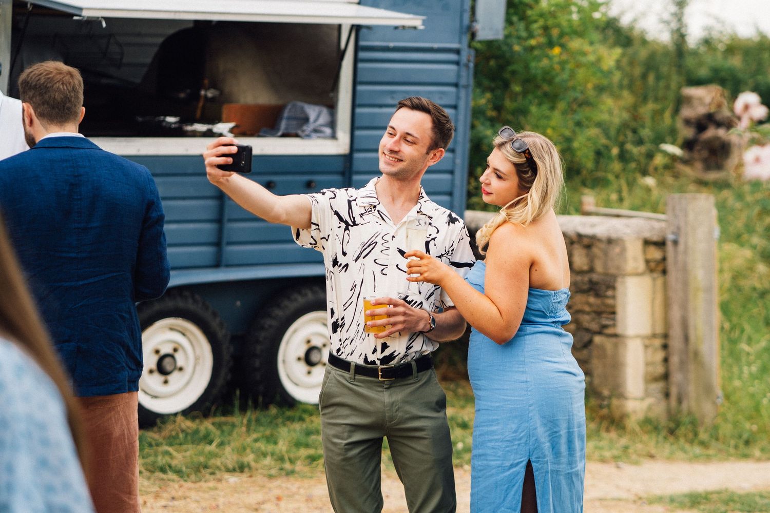 Guests enjoy drinks and conversation at an outdoor wedding reception near a rustic building.