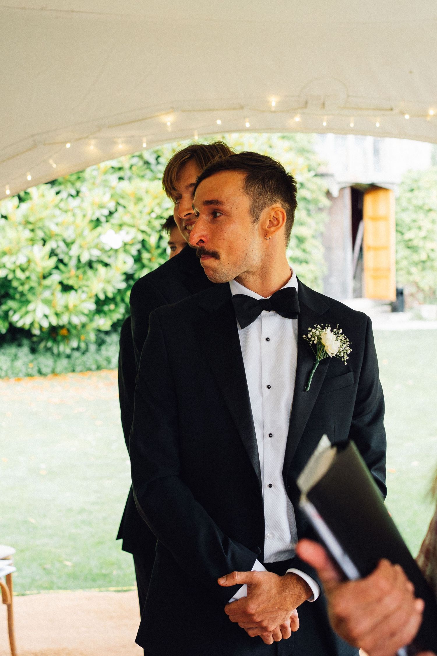 Groomsmen in black tuxedos and bow ties share an emotional moment under a tent during a wedding ceremony.
