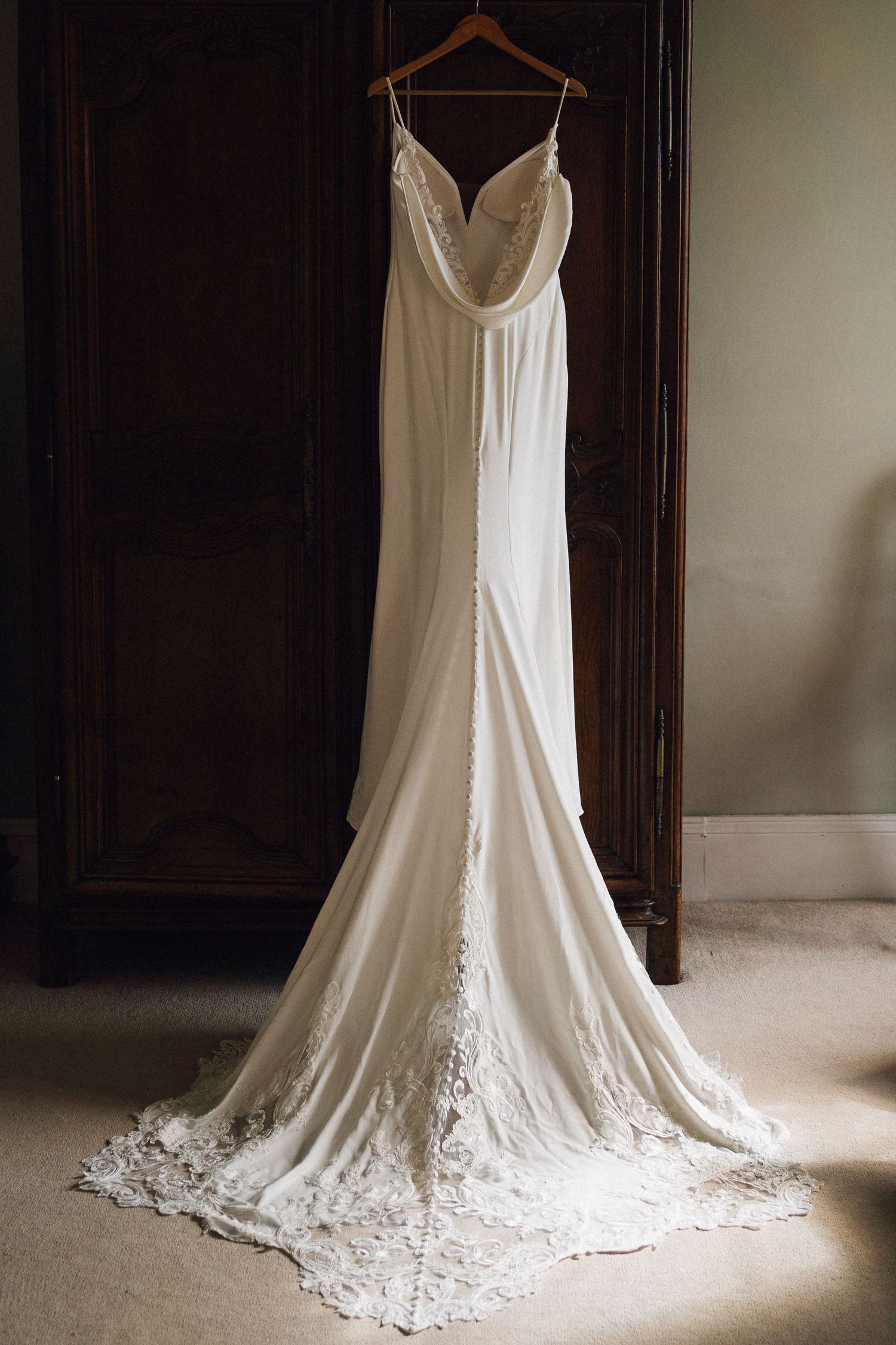 Elegant white wedding dress with lace train displayed in natural window light against dark wood panels.