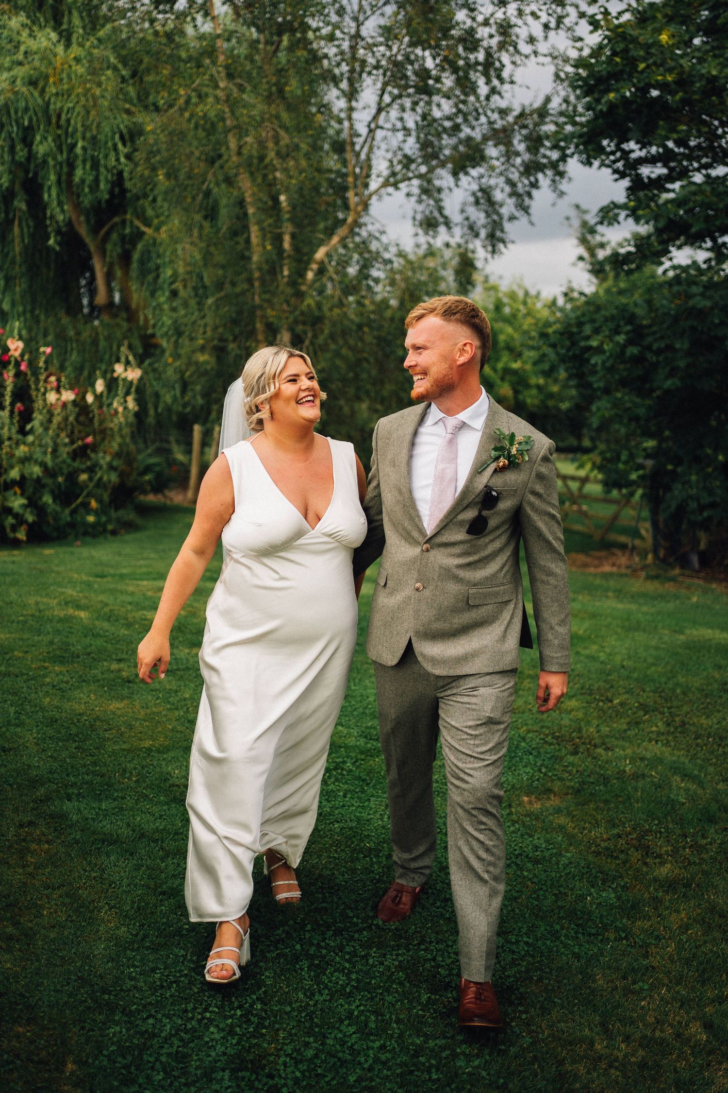 A couple in wedding attire walks together and laughs on a lush green lawn with willow trees in the background.