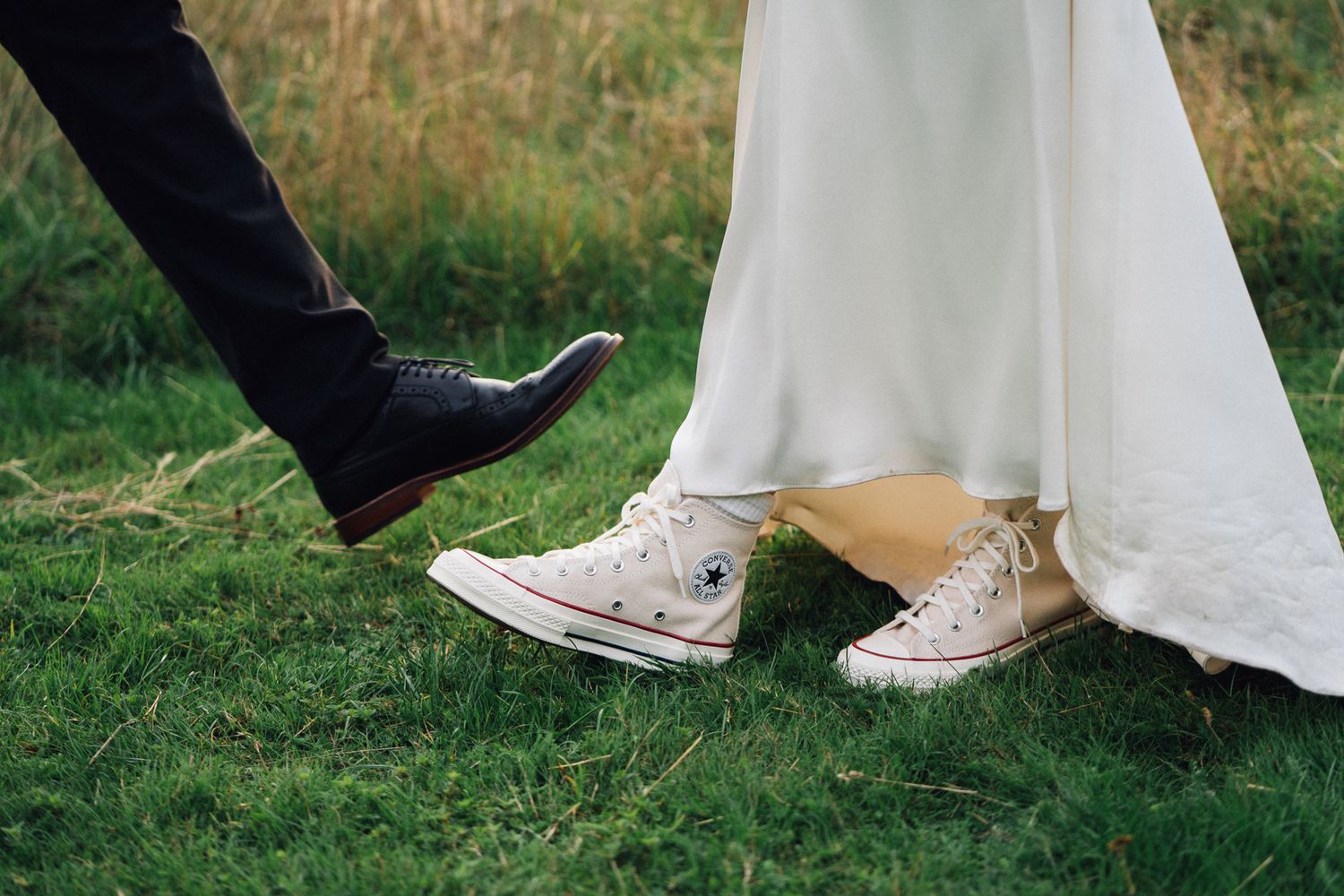 A playful wedding photo showing dress shoes and white Converse sneakers under a wedding dress on grass.