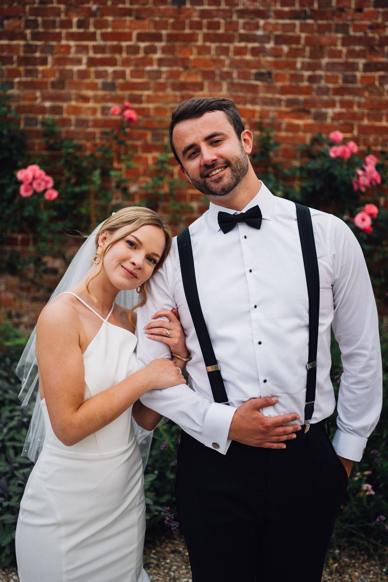 Romantic wedding couple poses against brick wall with pink roses in garden setting.