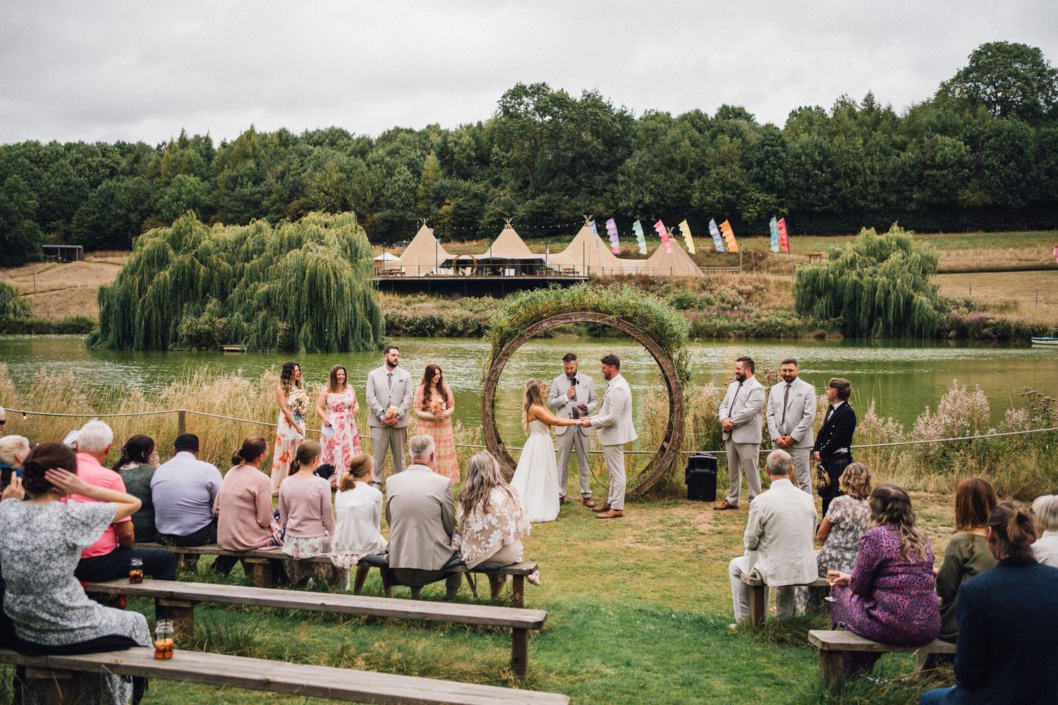 Outdoor wedding ceremony under a floral arch with bell tents and willow trees in the background.