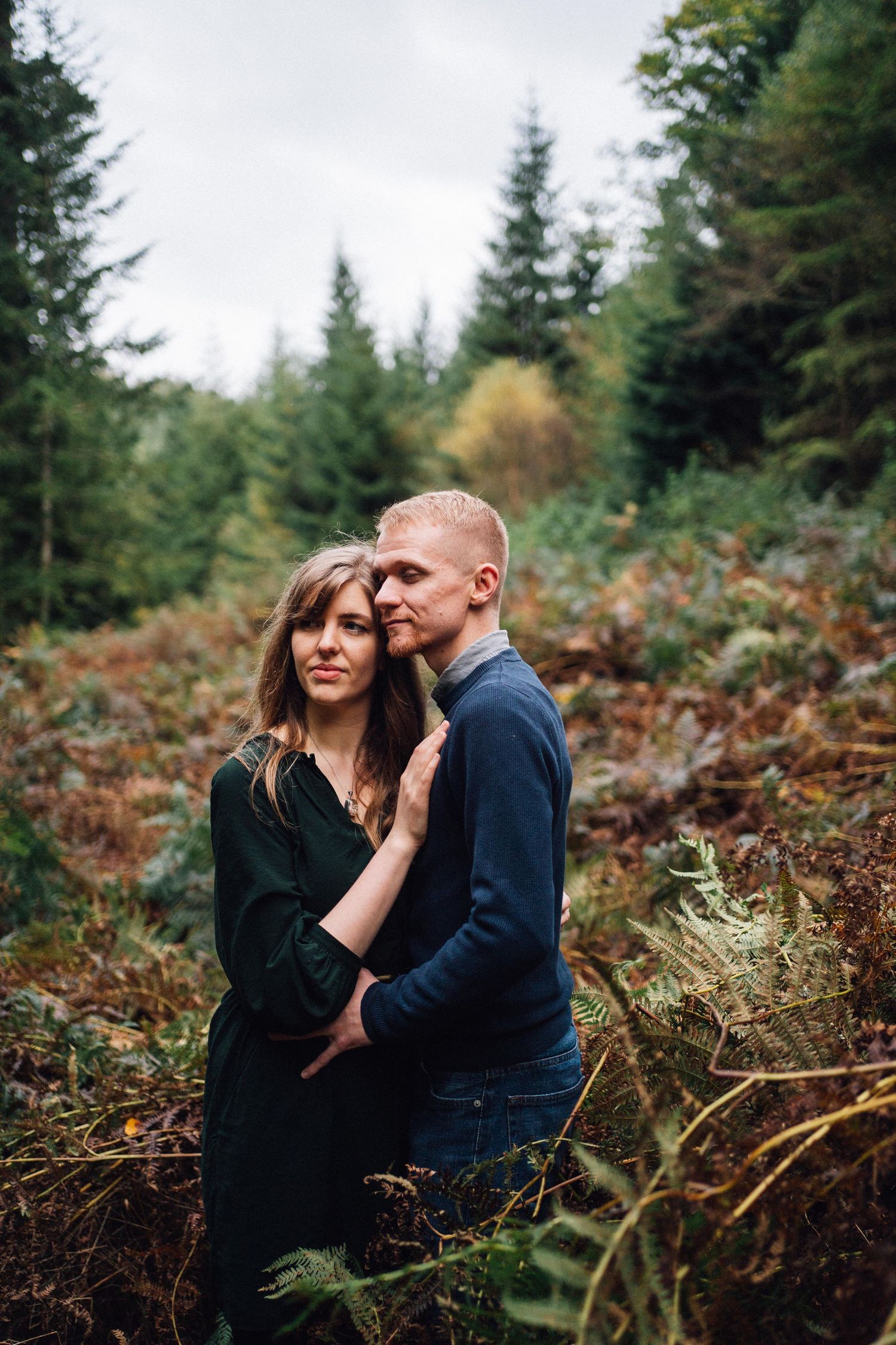 A romantic couple shares an intimate moment among ferns and evergreen trees in a forest setting.
