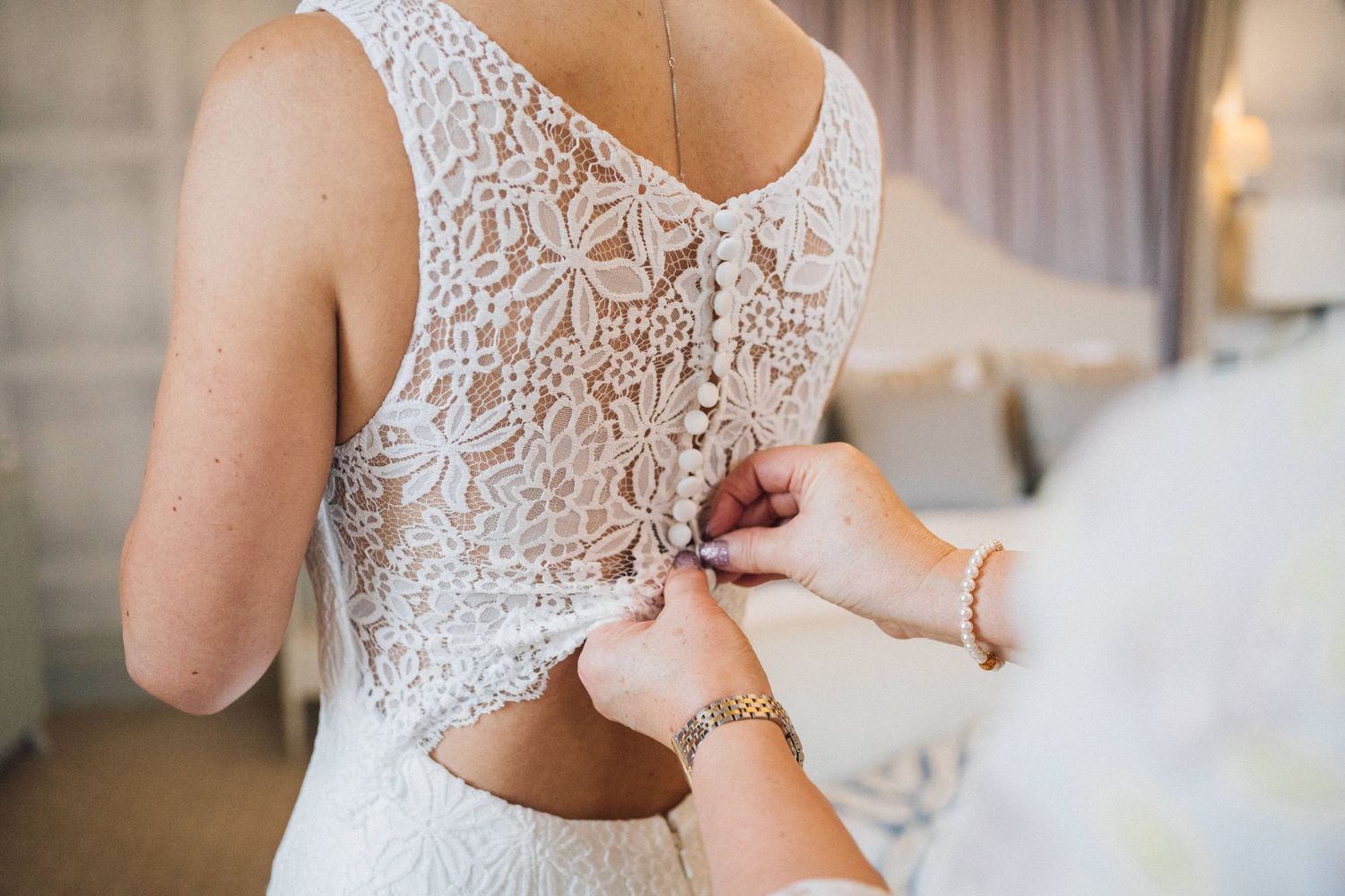 Close-up details of an elegant lace wedding dress being fastened in a brightly lit room.