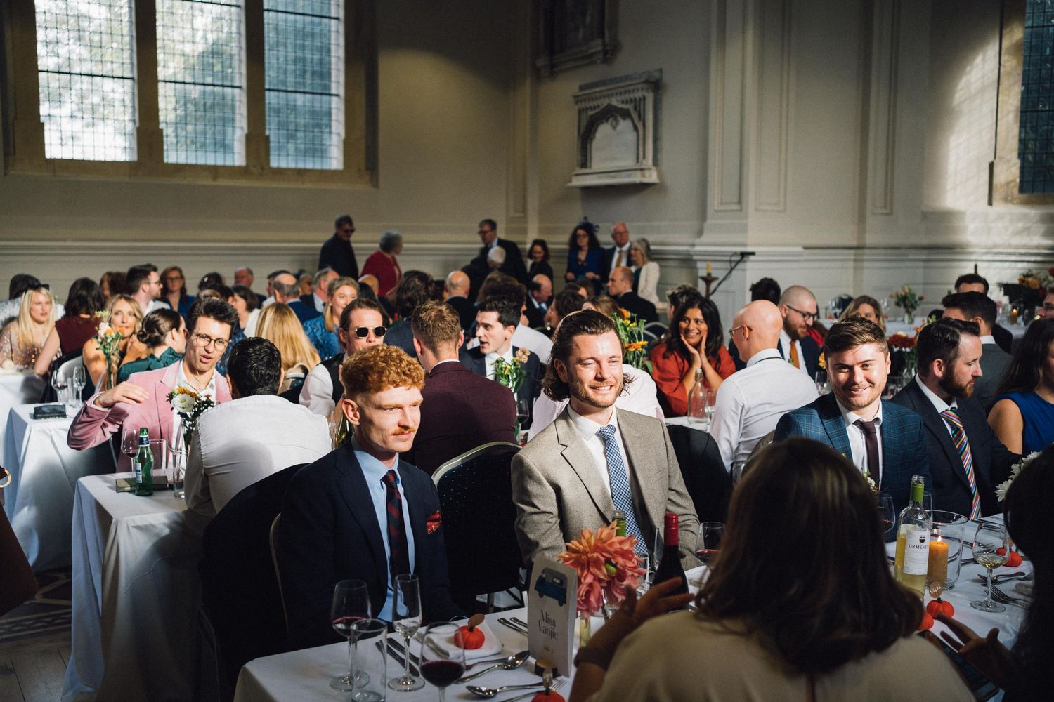 Large indoor wedding reception with guests dining at multiple decorated tables.
