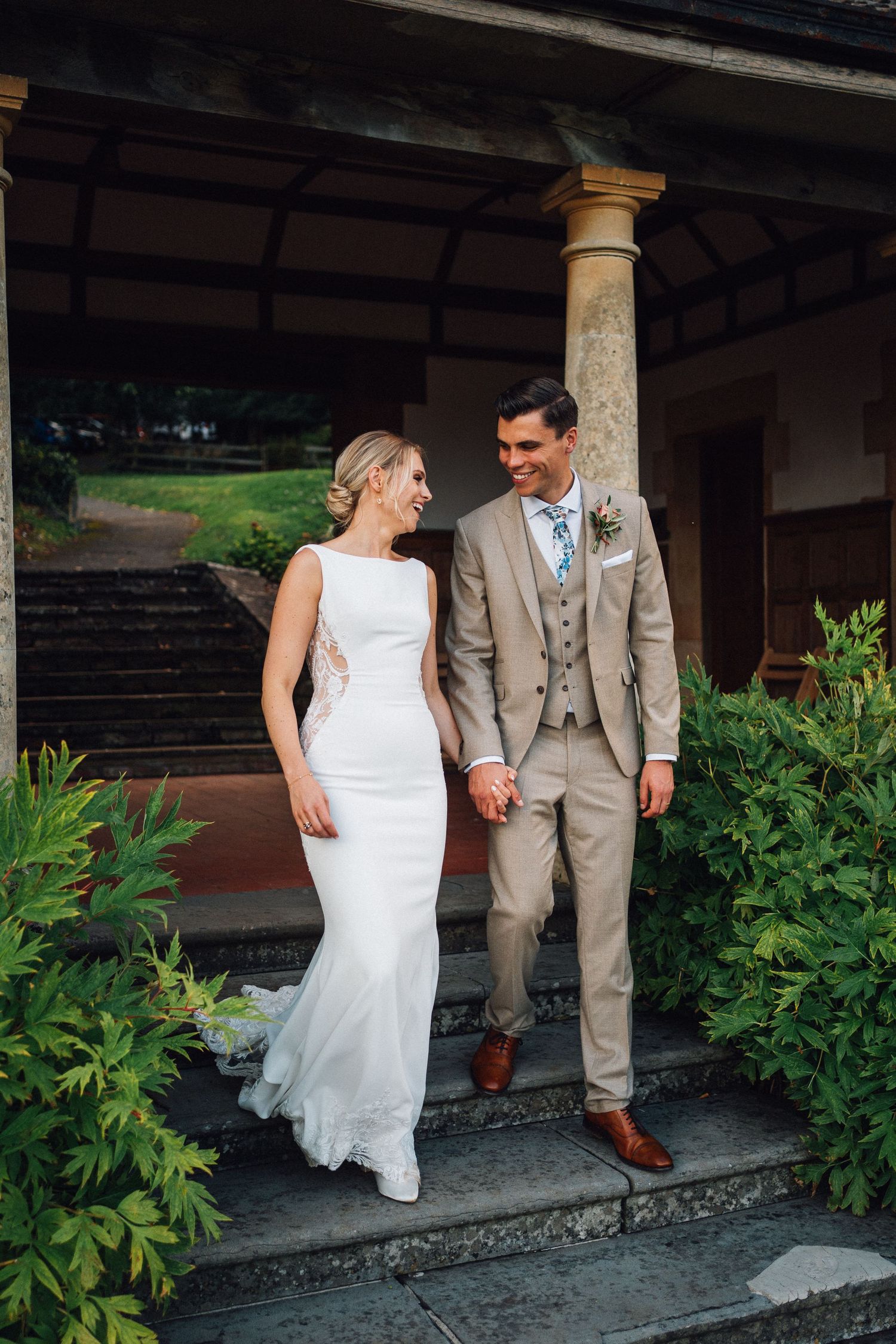 Couple in wedding attire walking along a stone path with greenery and columns.