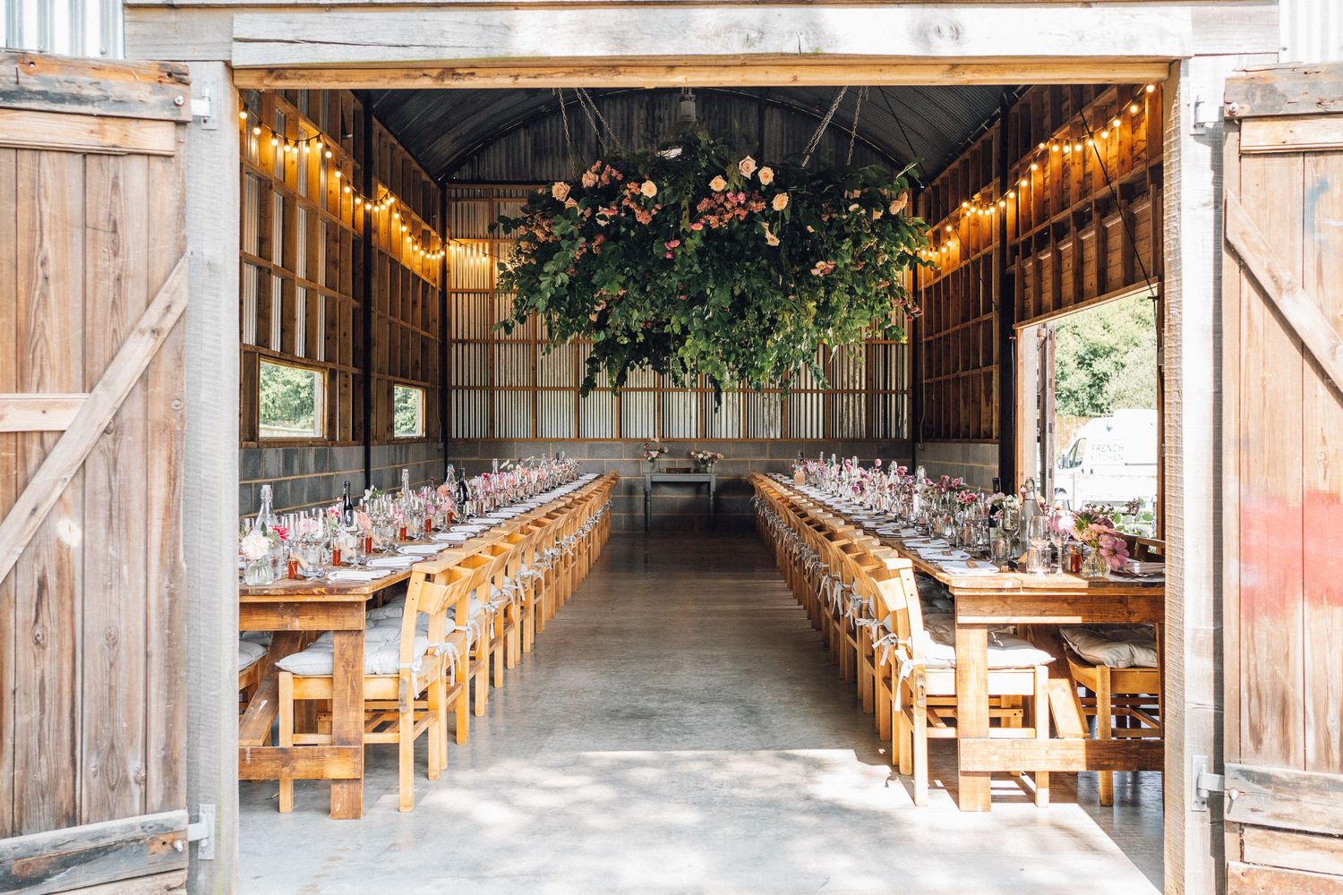 Rustic barn wedding reception with long wooden tables, string lights, and a lush floral ceiling installation.