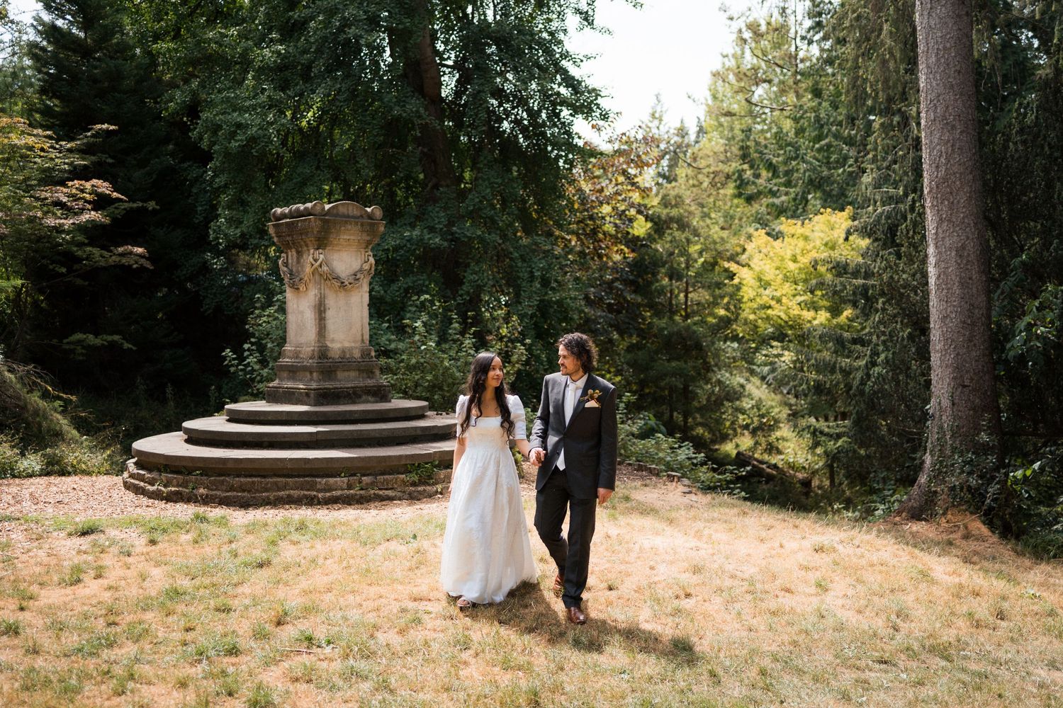 Bride and groom walking hand in hand through a scenic garden park with tall trees and stone monument.