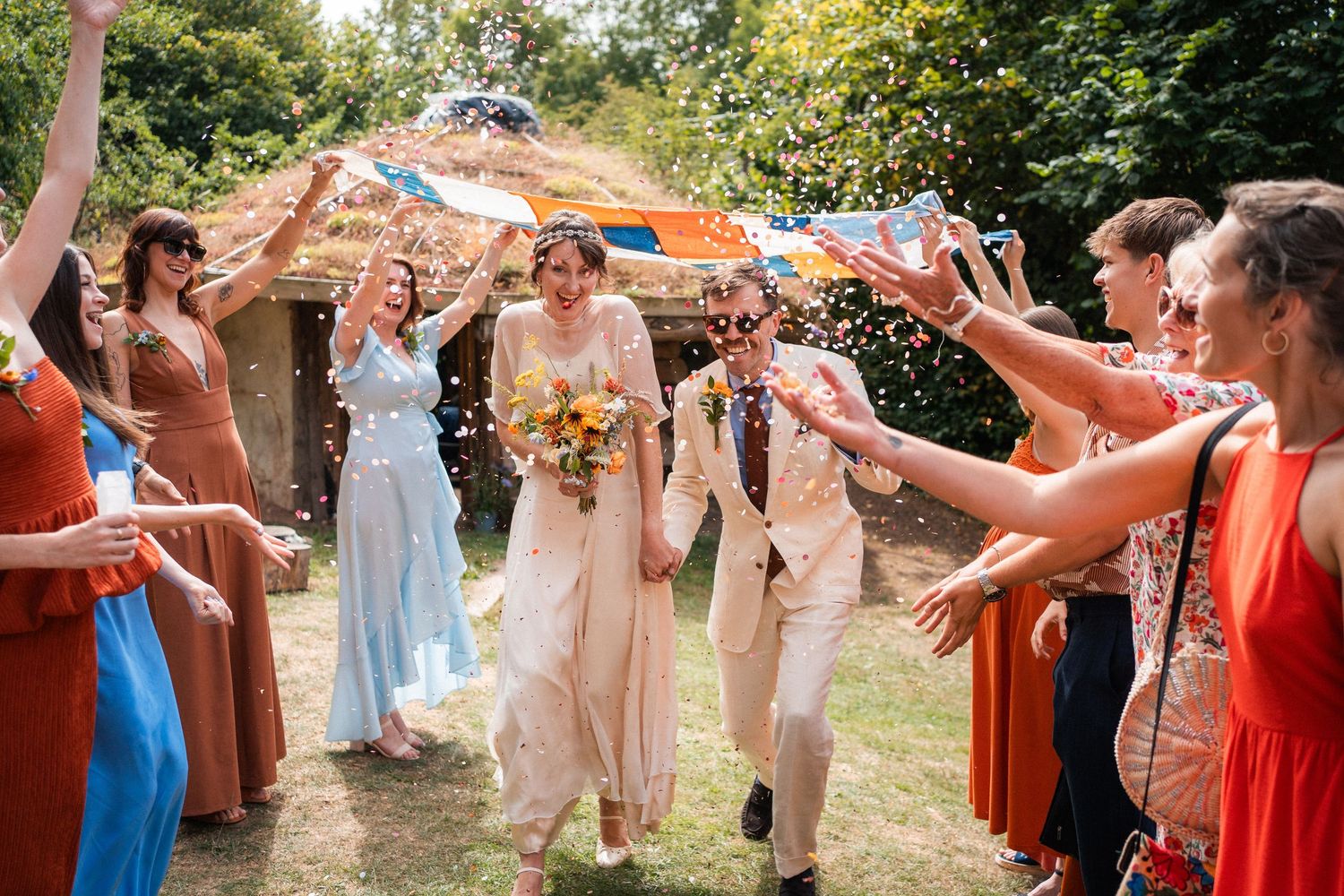 Bride and groom walk through confetti tunnel with colorful bridesmaids at outdoor rustic wedding celebration.