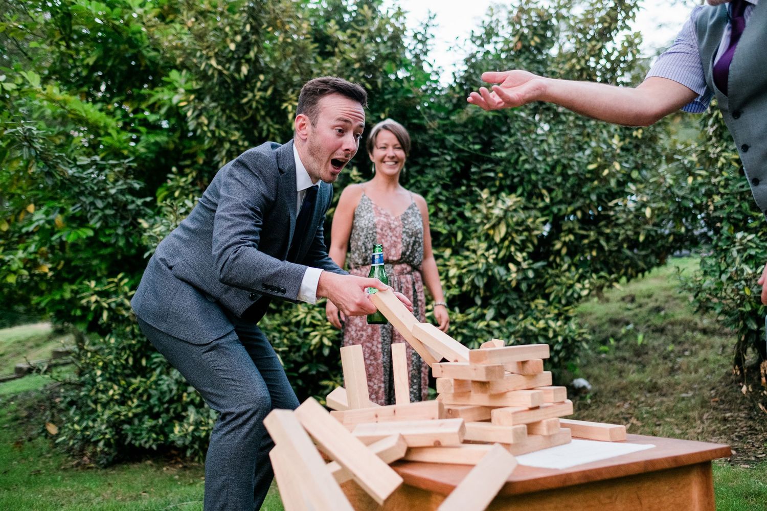 Wedding guests react as giant Jenga blocks fall during outdoor reception entertainment.