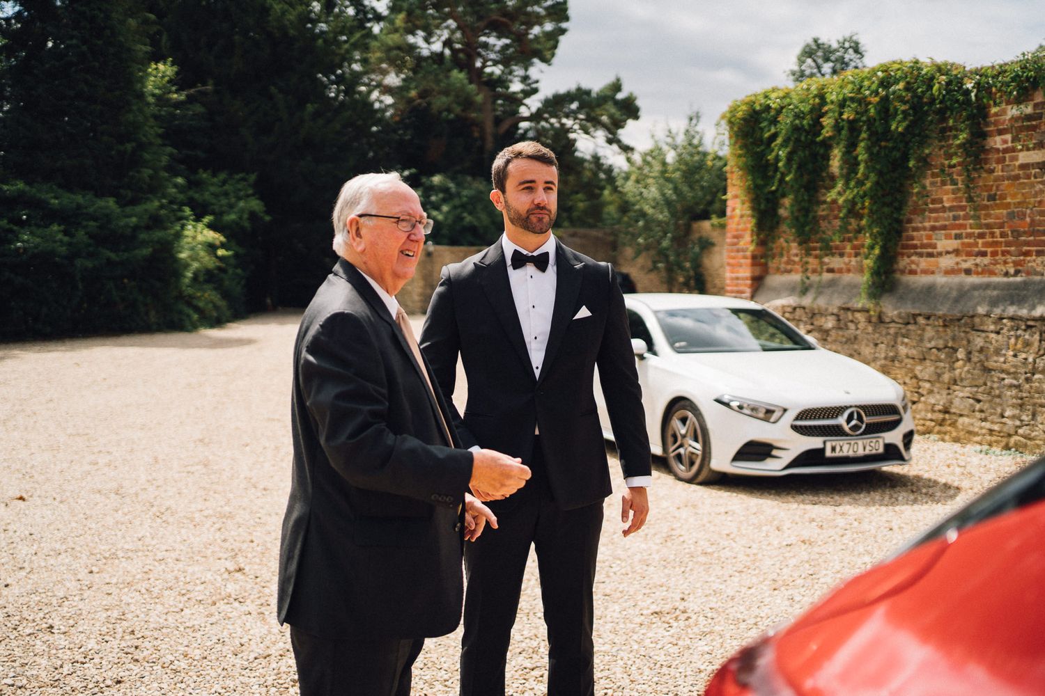 Group of men in tuxedos gathered around luxury vehicles in a stone courtyard.