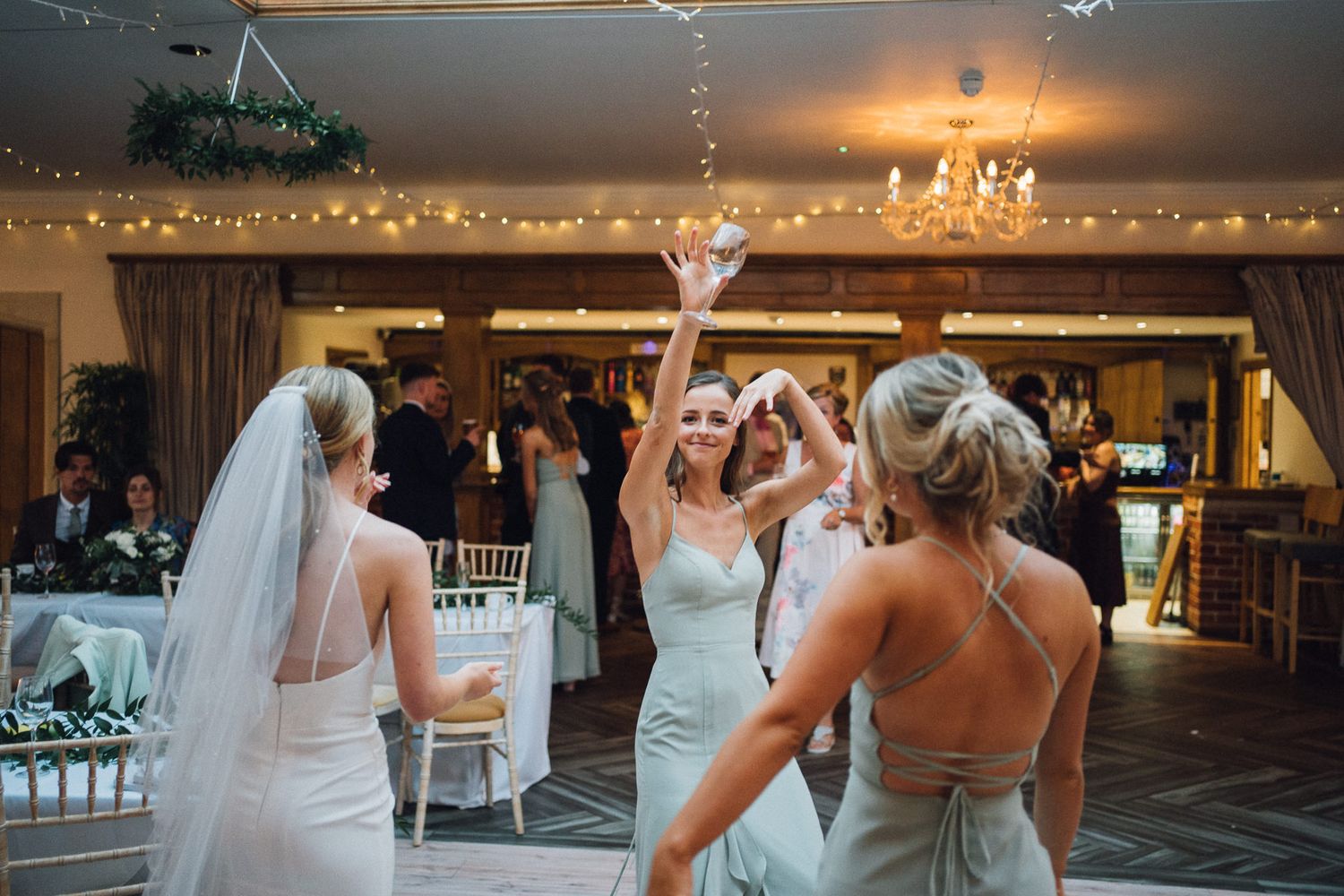 Wedding guests dancing and celebrating in a reception hall with twinkling lights and chandeliers.