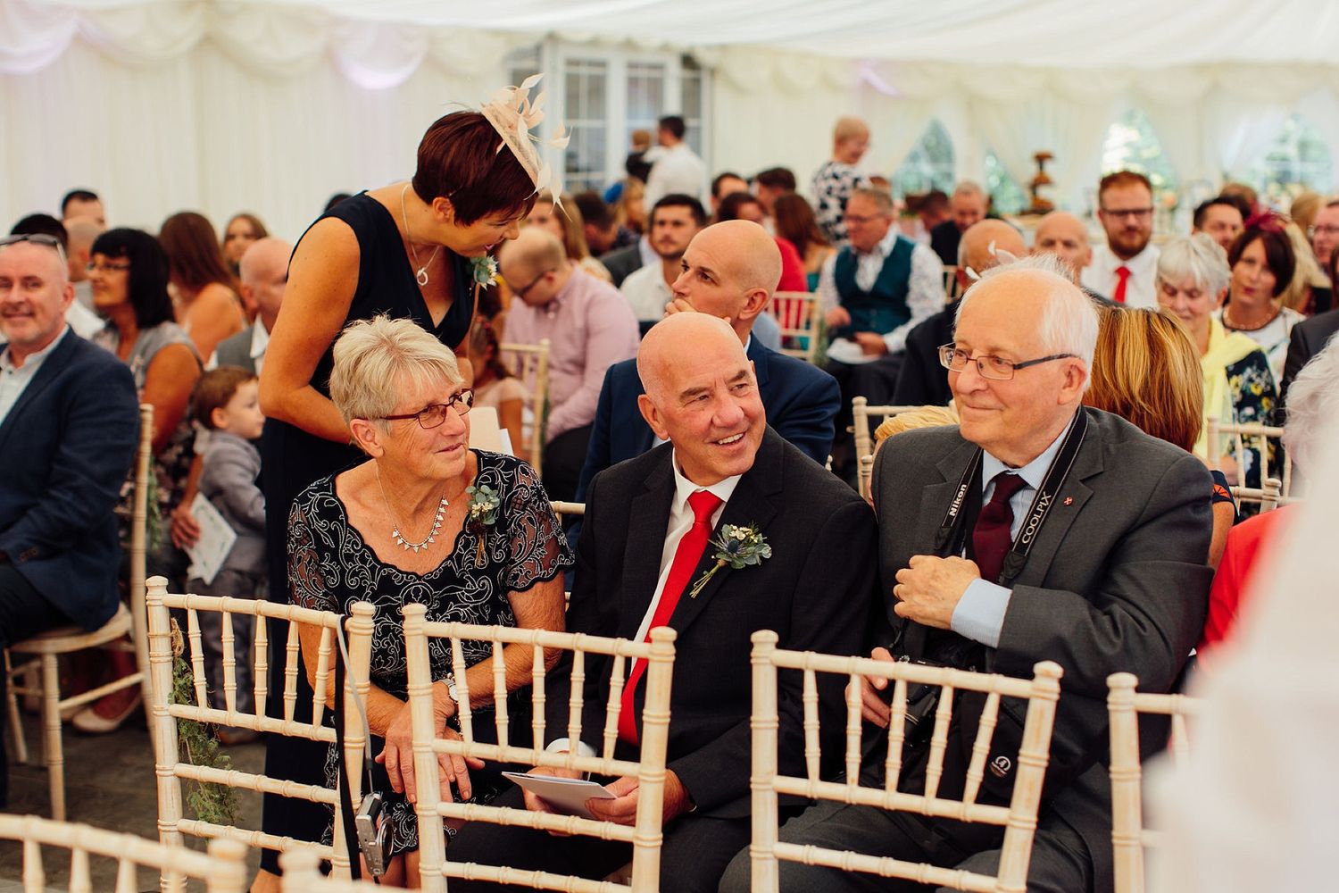 Wedding guests seated in white chiavari chairs under an elegant white tent during ceremony.