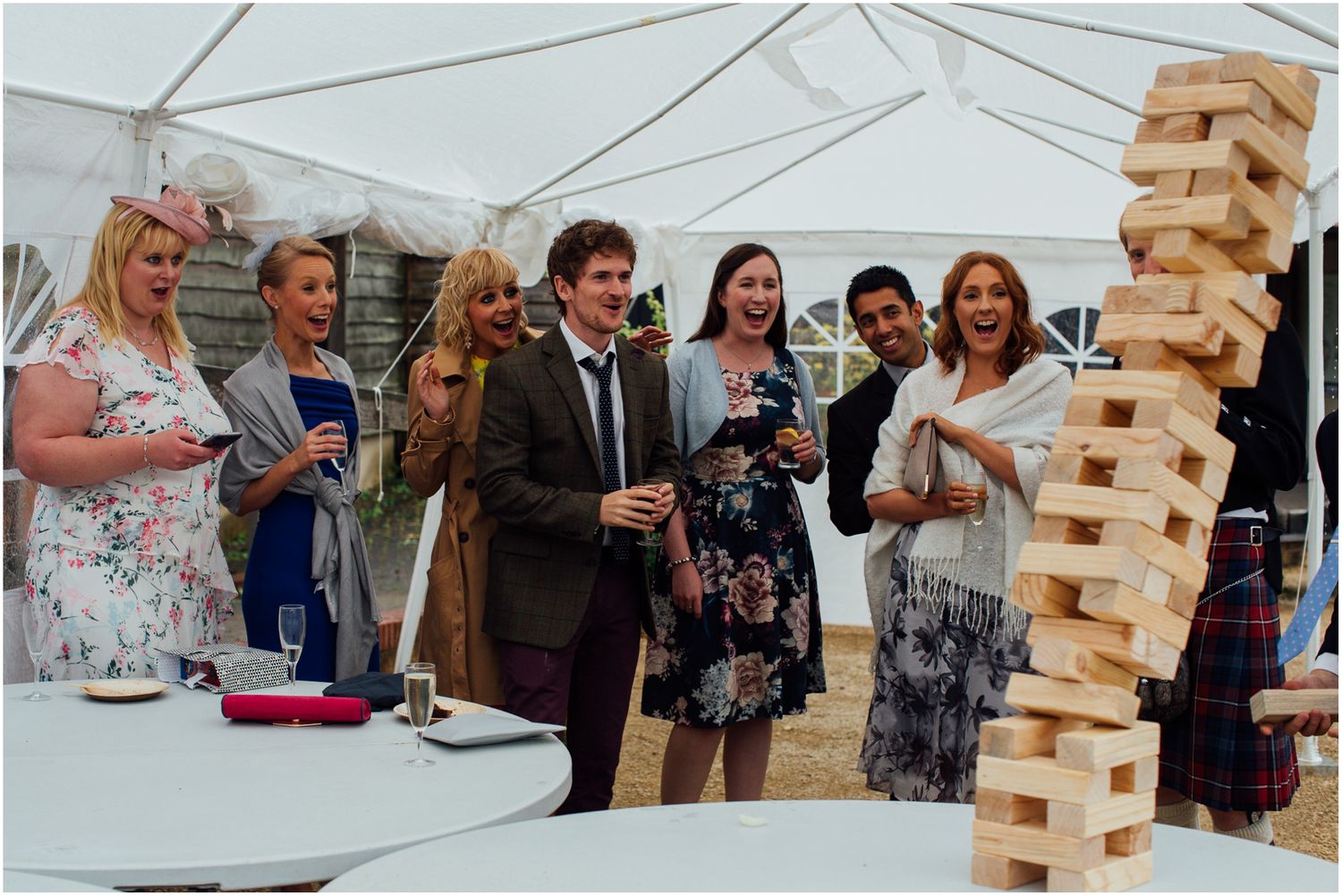 Large crowd watching intense giant Jenga game at outdoor party under white tent.