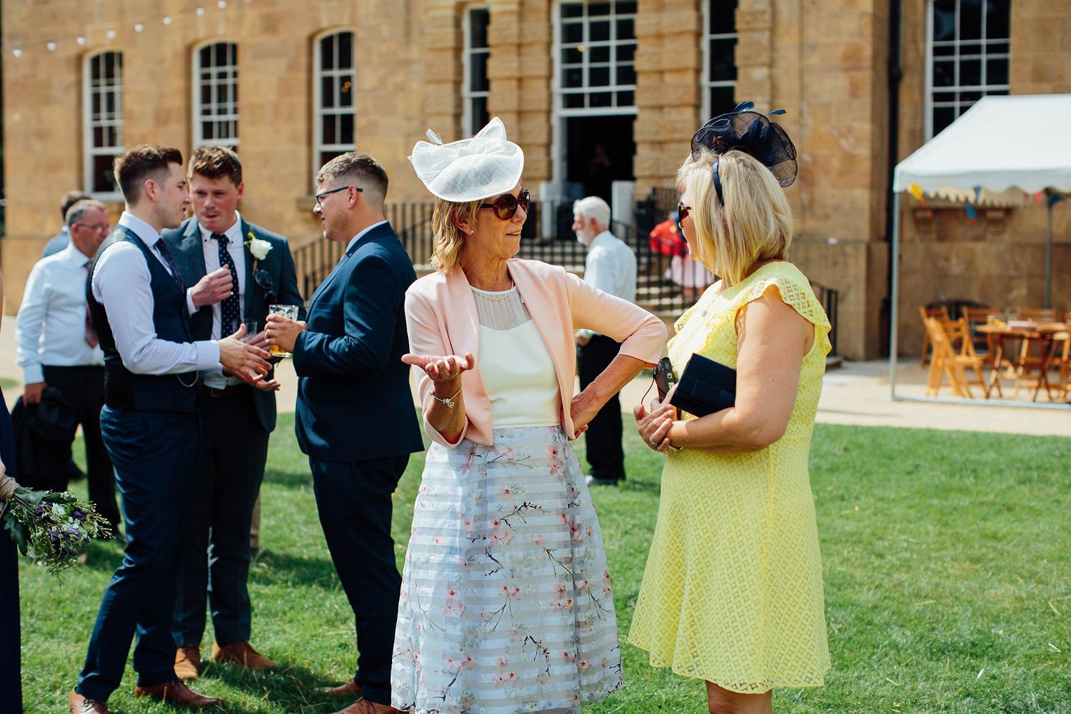 Wedding guests mingling on lawn outside elegant stone building during summer reception.