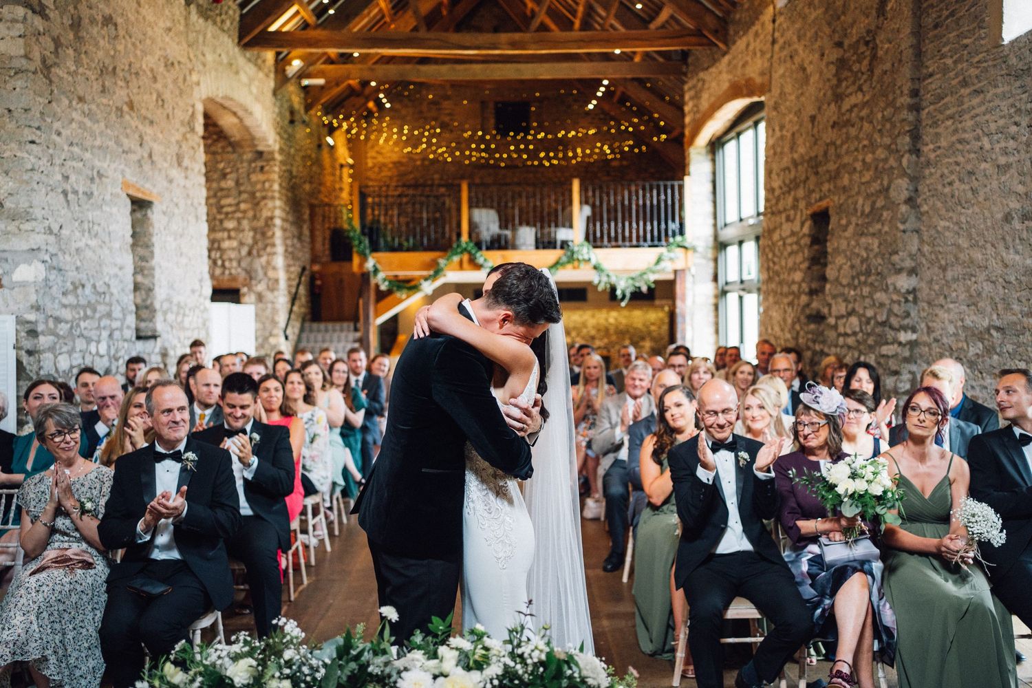 Wedding guests celebrate during a rustic barn reception with string lights and exposed stone walls.