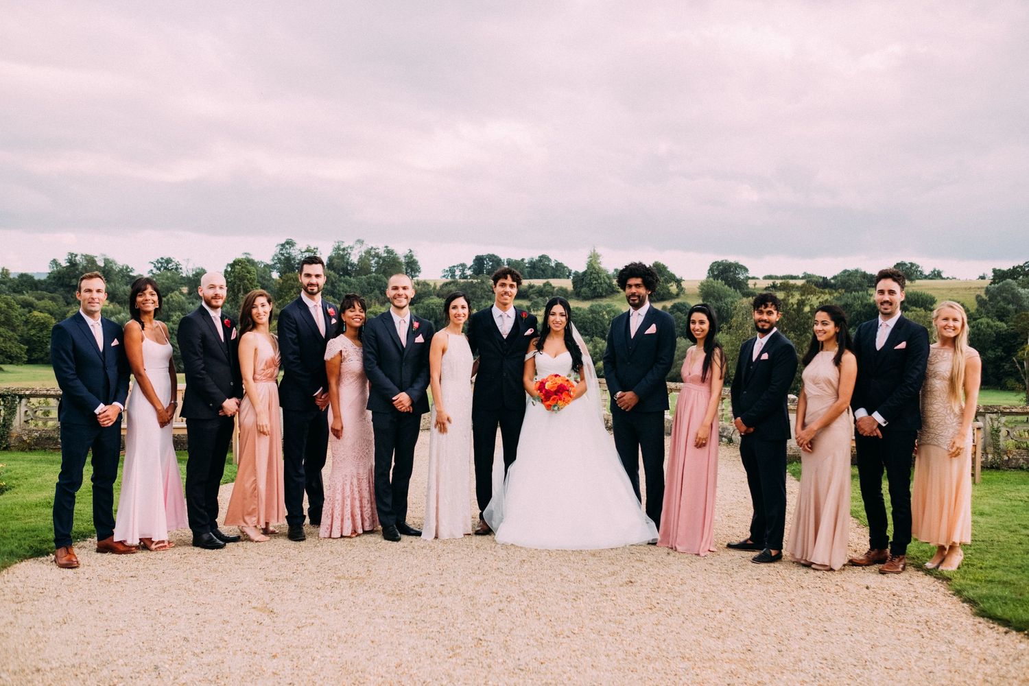 Wedding party in formal attire lined up for photos on a grassy lawn under cloudy skies.