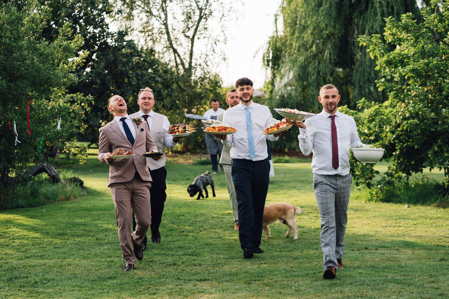 A sequence of wedding party members walking together through a lush green garden setting at sunset.