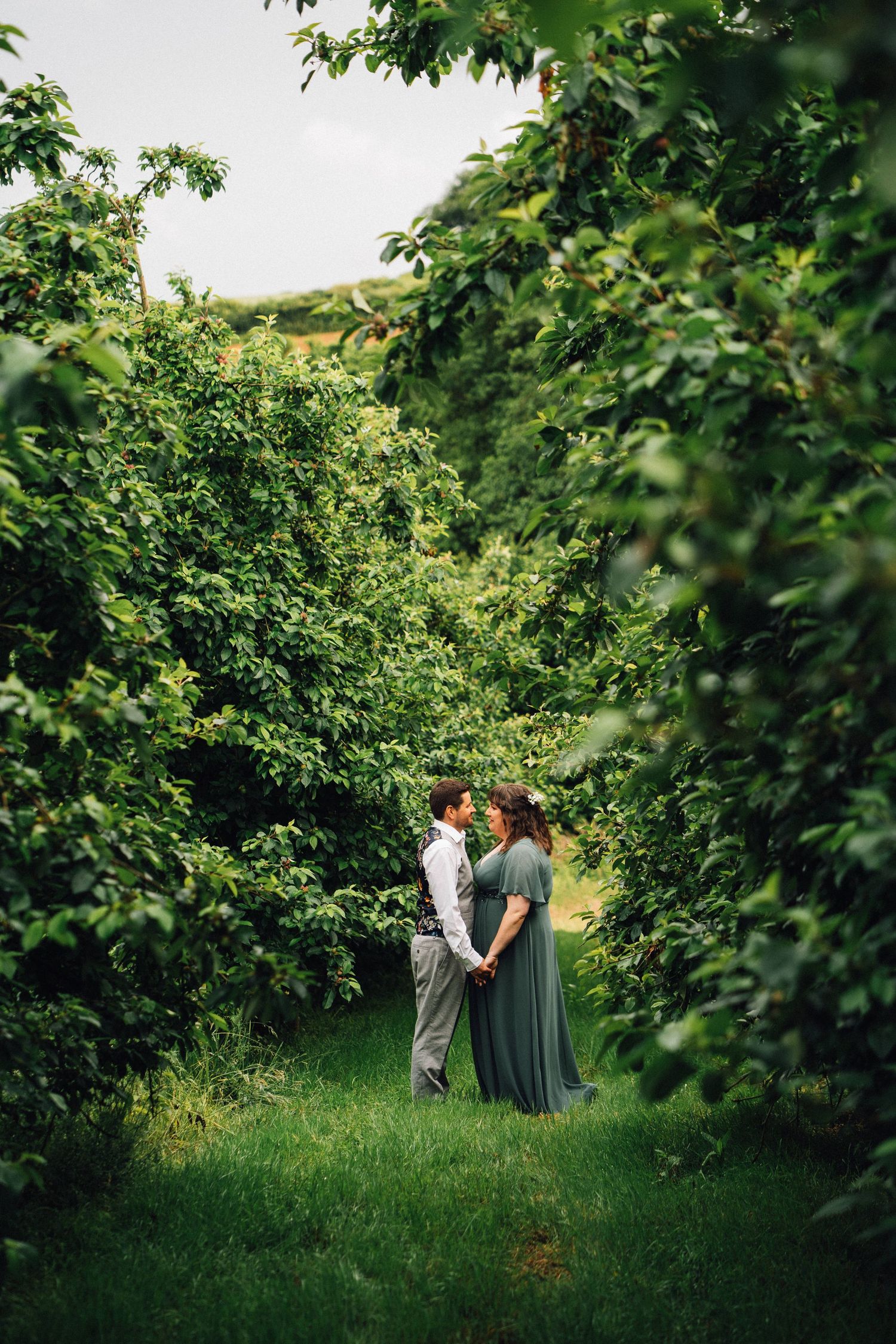 Two people walking through a lush green garden path surrounded by tall hedges on a sunny day.