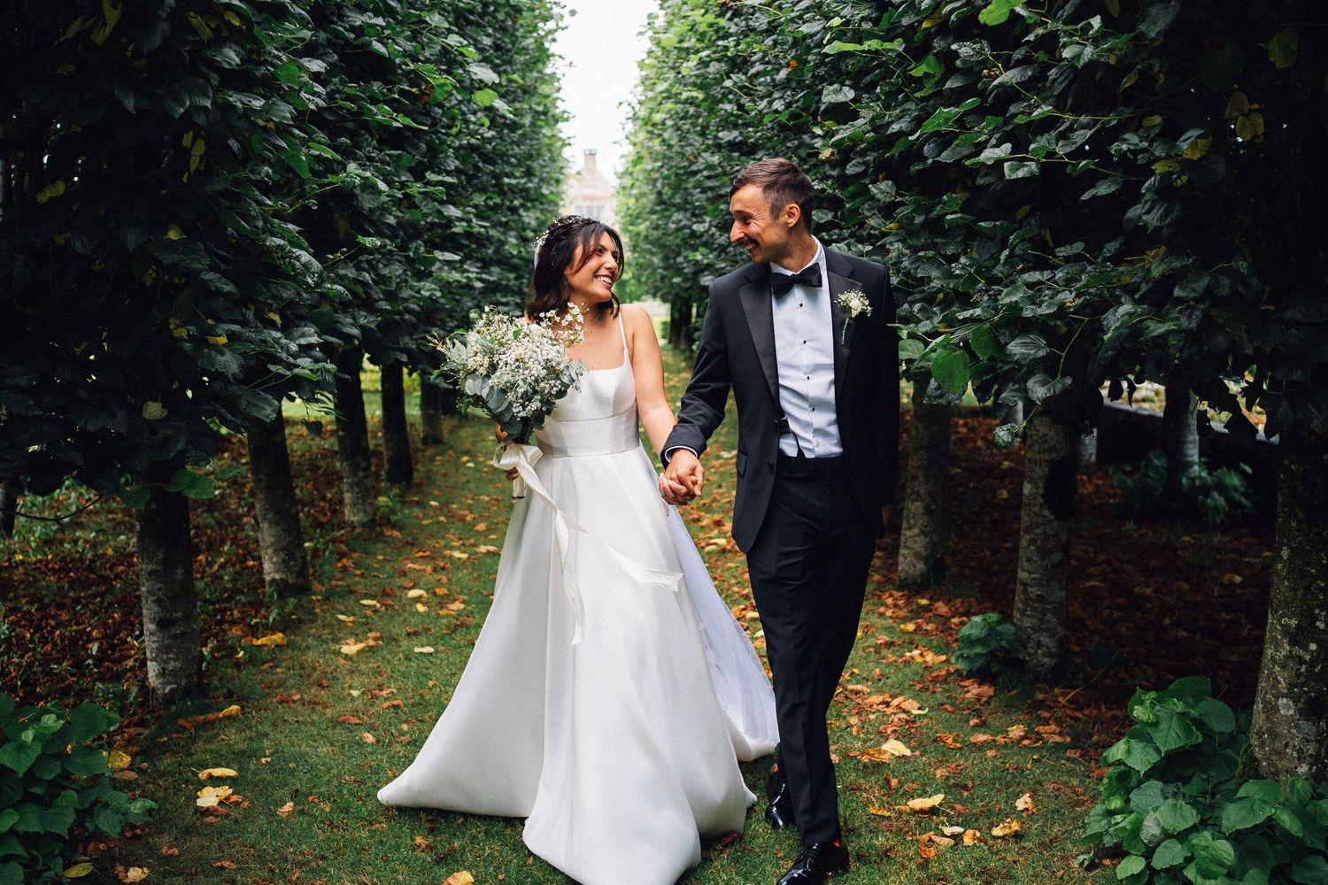 Bride and groom walking hand in hand through a lush green tree-lined garden path on their wedding day.