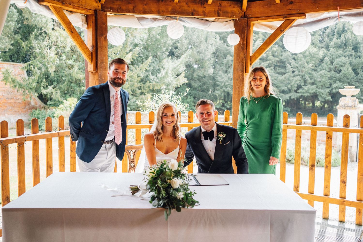 Wedding party gathers under a rustic wooden pavilion surrounded by trees during an outdoor celebration.