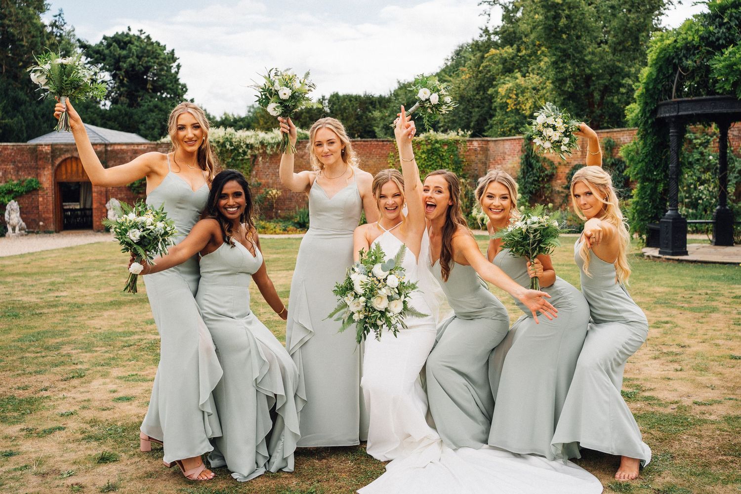 Bridesmaids in light gray dresses pose playfully with bride in outdoor garden setting.