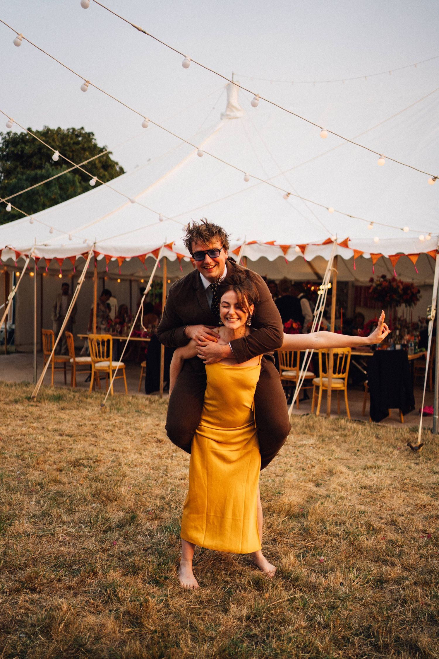 A couple celebrates at an outdoor wedding reception under a tent with string lights at dusk in Somerset