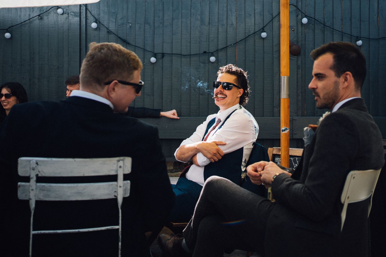 Three professionals having an outdoor business conversation while seated in chairs against a dark wall with string lights.