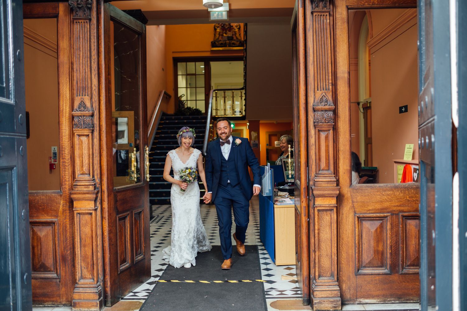 A couple exits through ornate wooden doors at their wedding venue with warm lighting and architectural details.