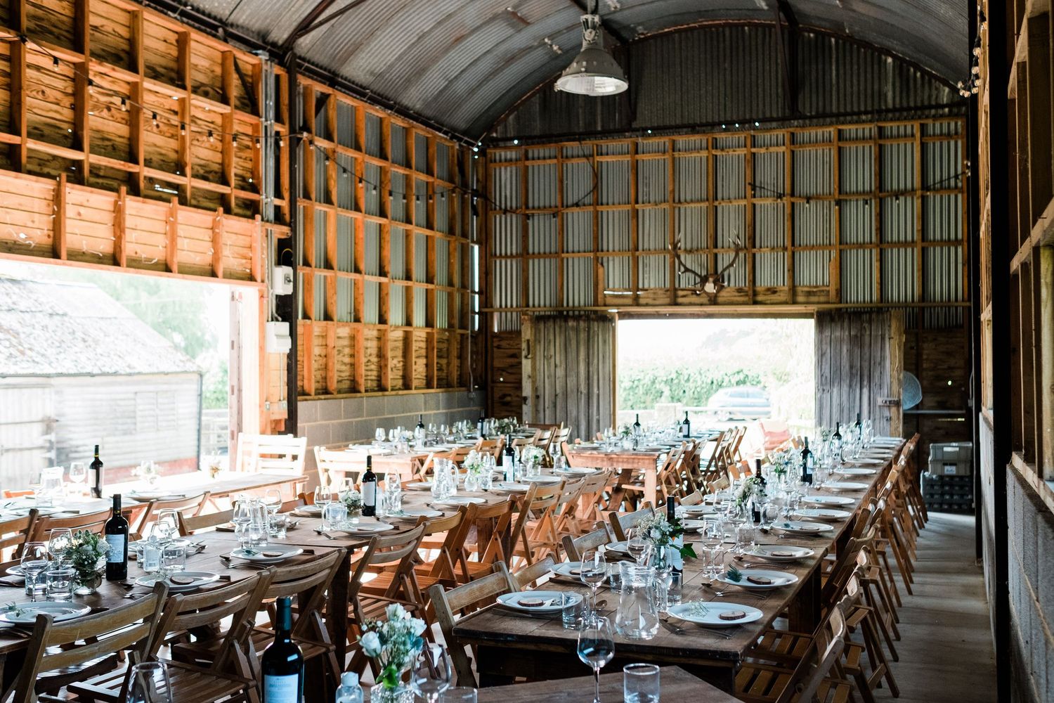 Rustic barn wedding reception venue with long wooden tables and chairs set up under exposed beams and natural lighting.