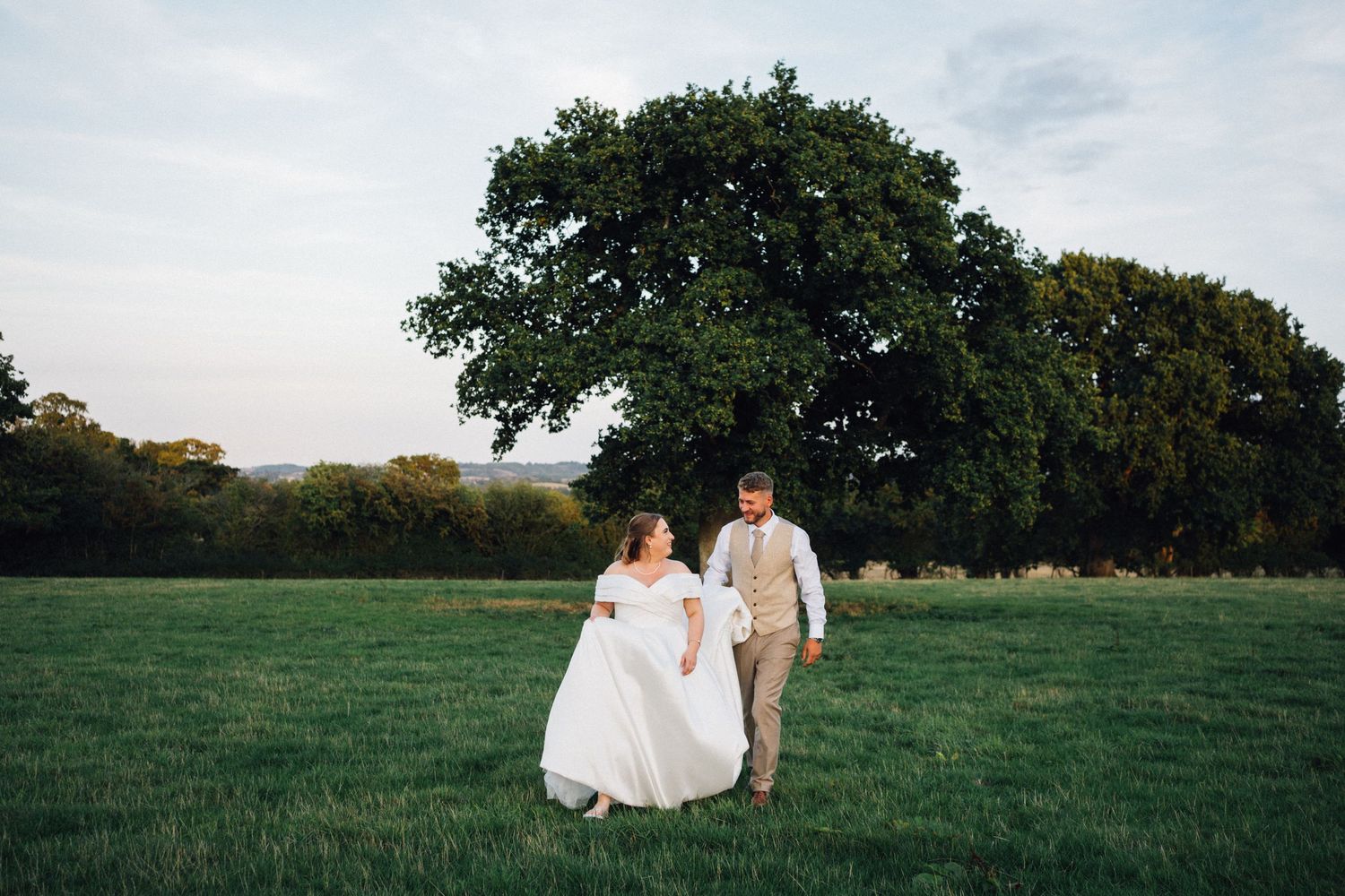A couple in wedding attire sits together on a grassy hillside during golden hour at sunset.