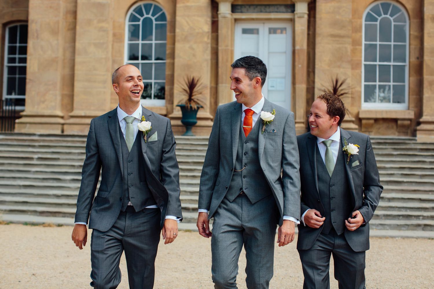 Three groomsmen in grey suits with orange ties laugh and walk together outside an elegant building on a wedding day.