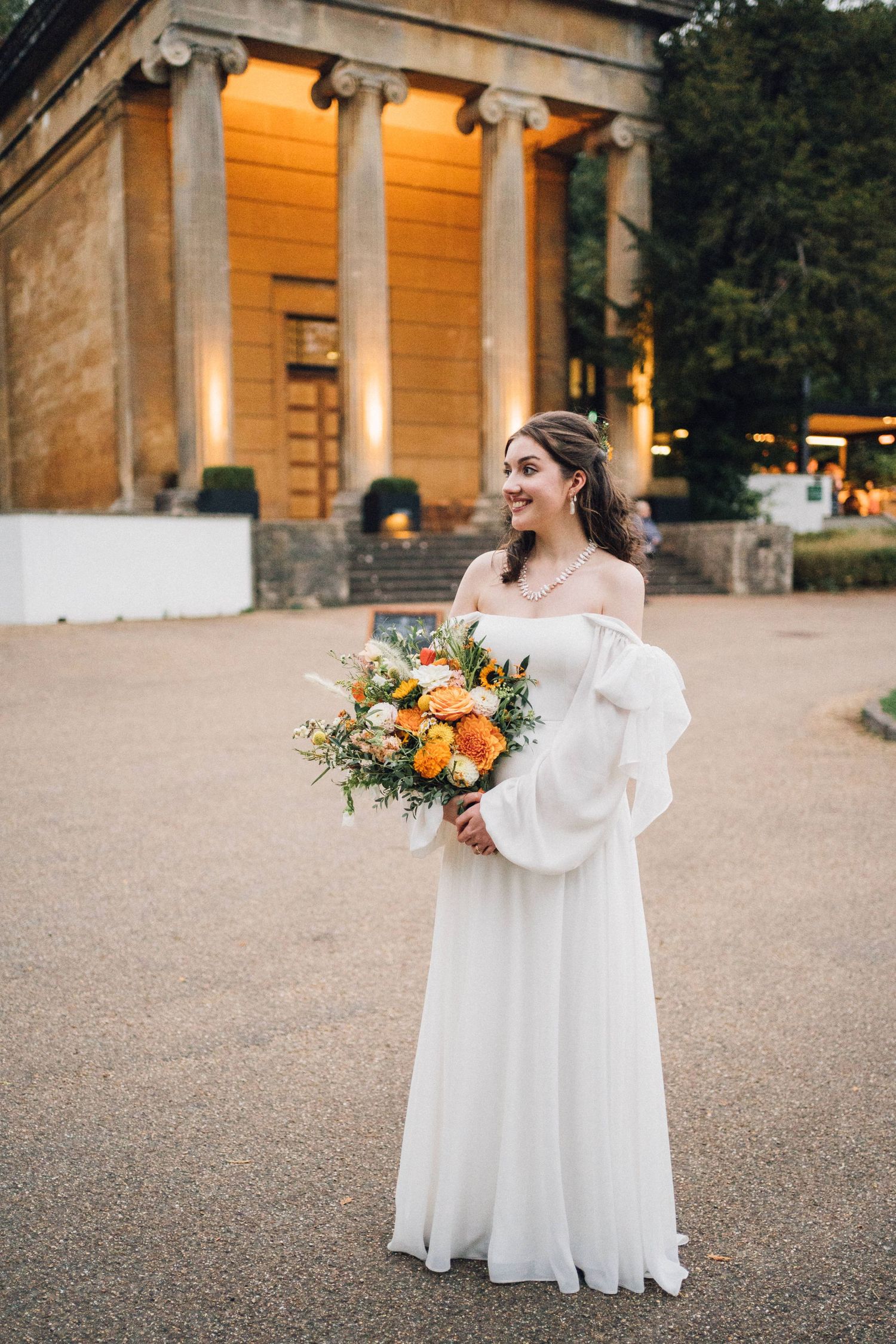 Smiling bride in white off-shoulder gown holding orange bouquet stands before illuminated classical building at dusk.