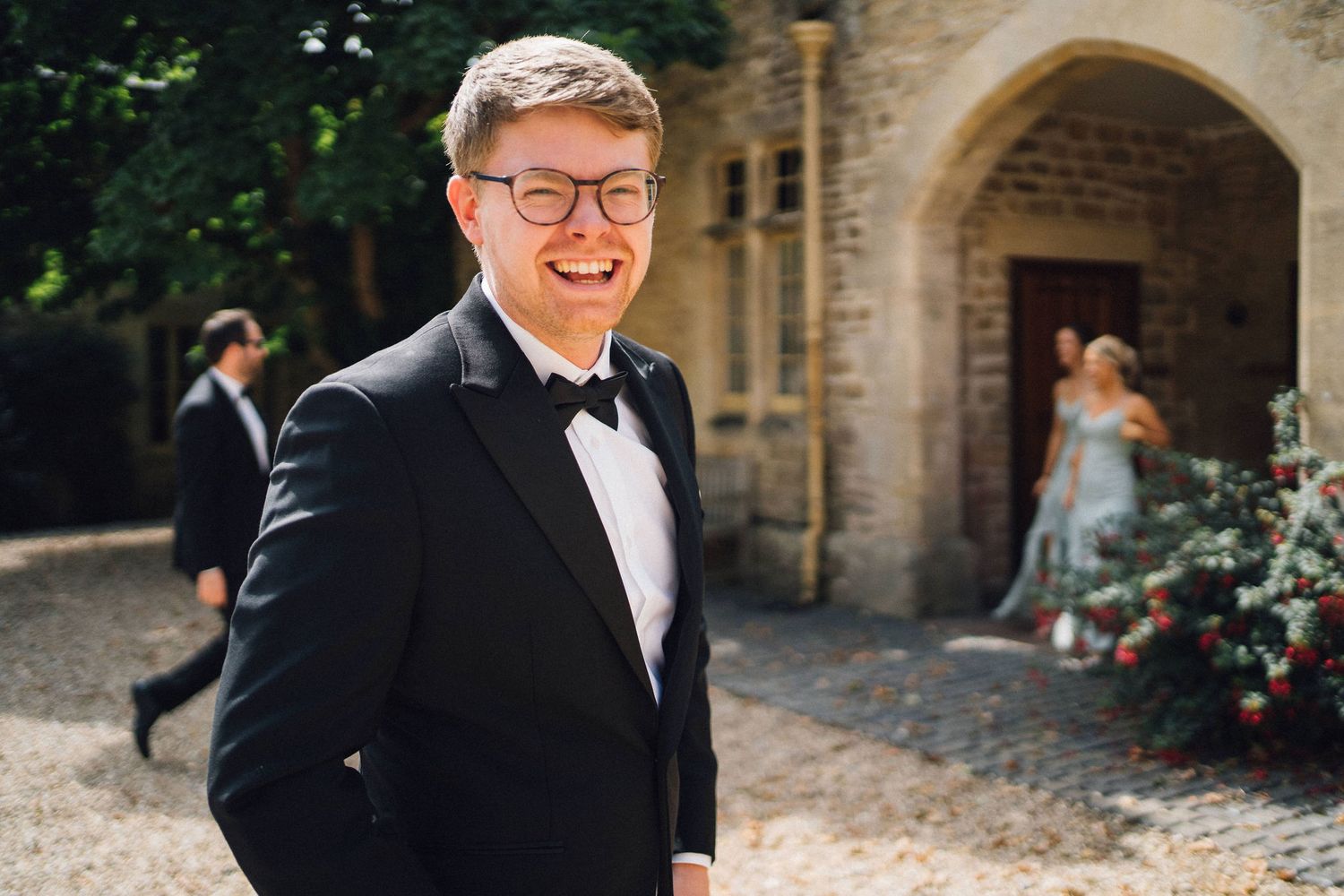 A person in a black tuxedo and bow tie smiles joyfully at a wedding venue with stone archways in the background.