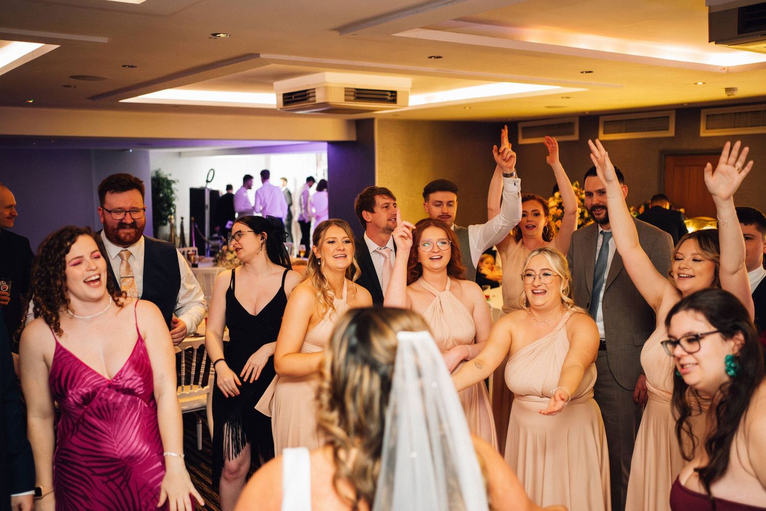 Wedding guests dance and celebrate with raised arms on a dimly lit indoor dance floor.