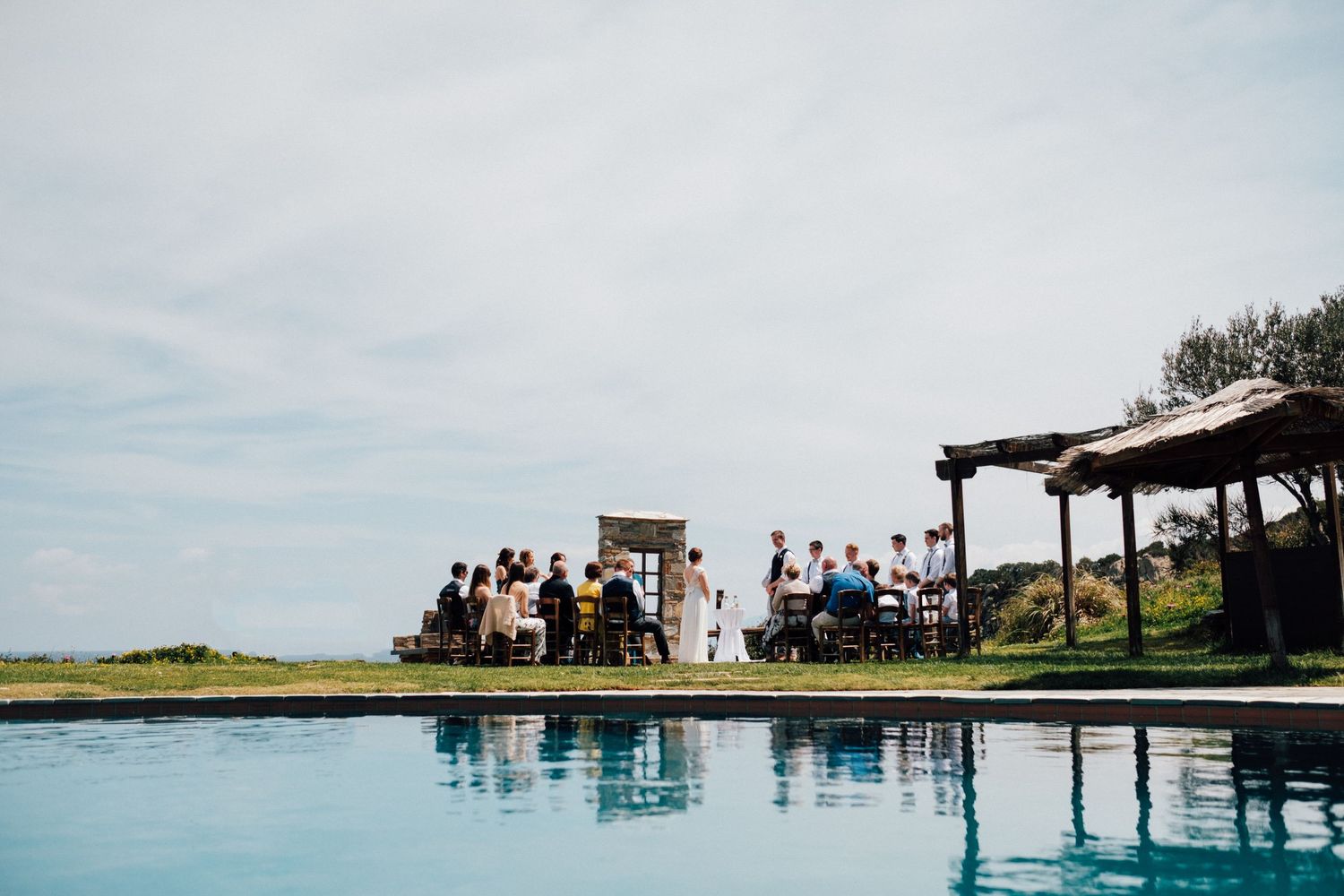Outdoor wedding ceremony space with guests gathered by infinity pool overlooking the ocean.