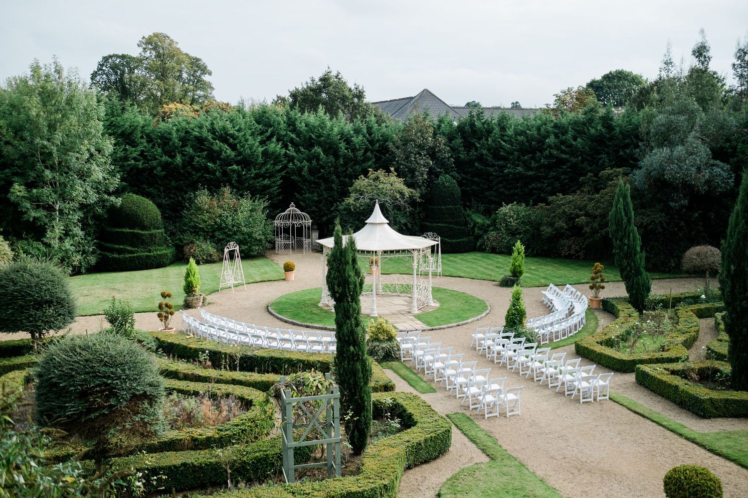 Elegant outdoor wedding ceremony setup with white chairs and a gazebo in a formal garden setting.
