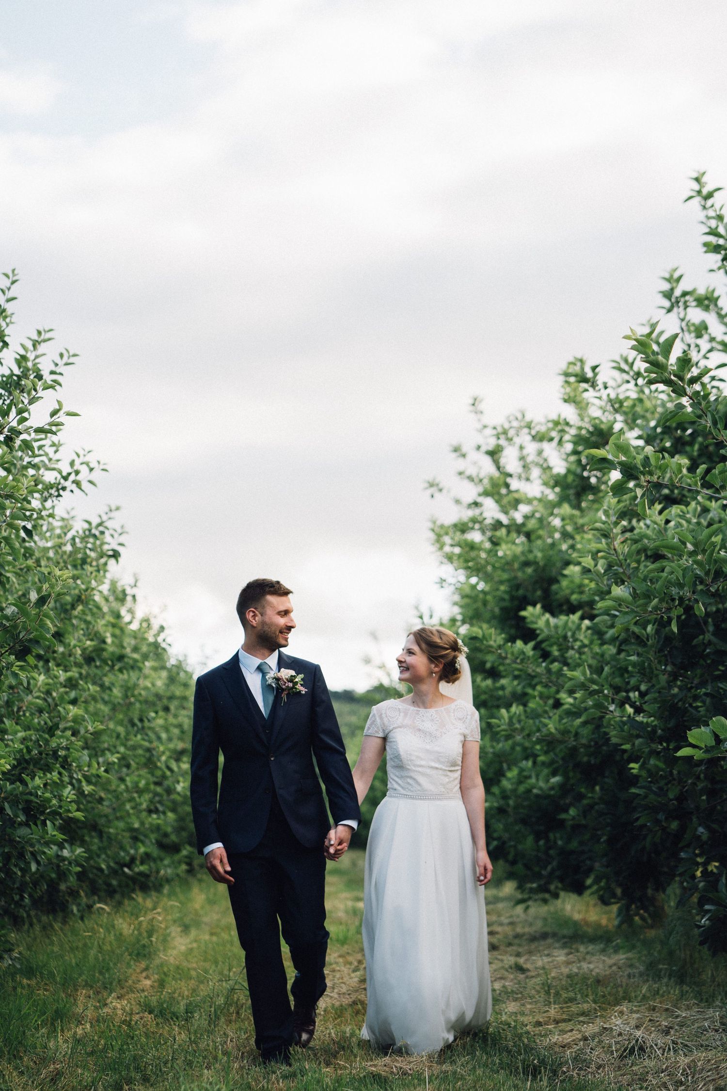 A romantic wedding portrait of a couple walking between rows of green trees.