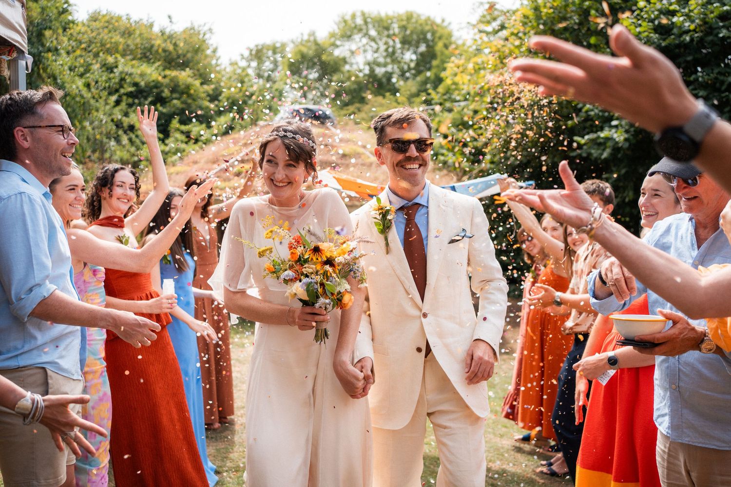 Happy newlyweds walk through colorful confetti tunnel with smiling guests at sunny outdoor wedding.