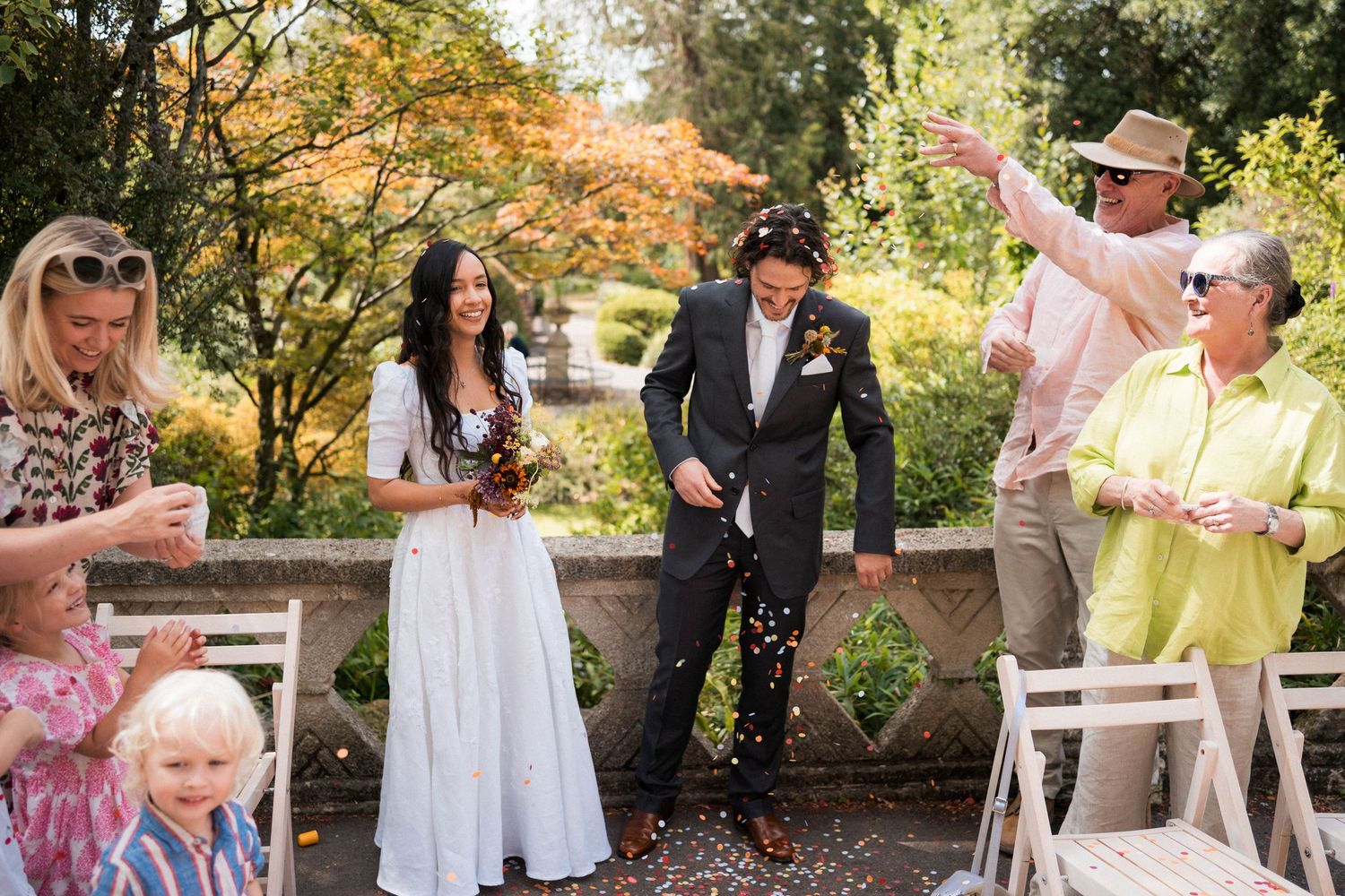 Newlywed couple walks down aisle as guests toss confetti outdoors in a garden wedding ceremony.