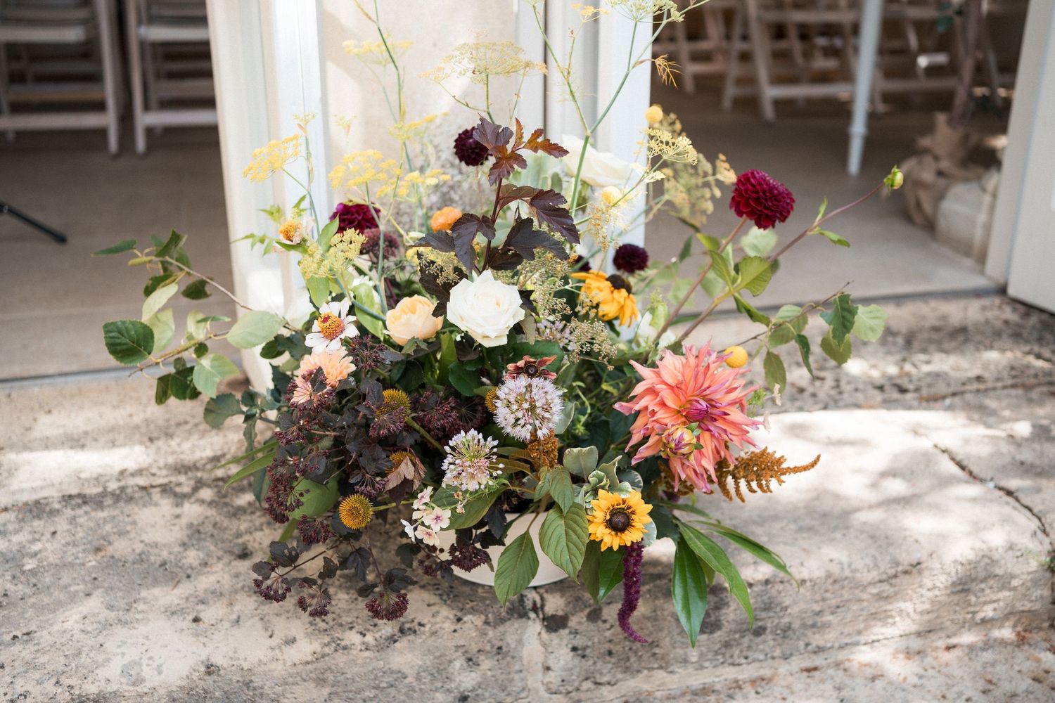 Lush garden floral arrangement with dahlias, sunflowers, and wildflowers in warm tones displayed on ground at outdoor wedding.